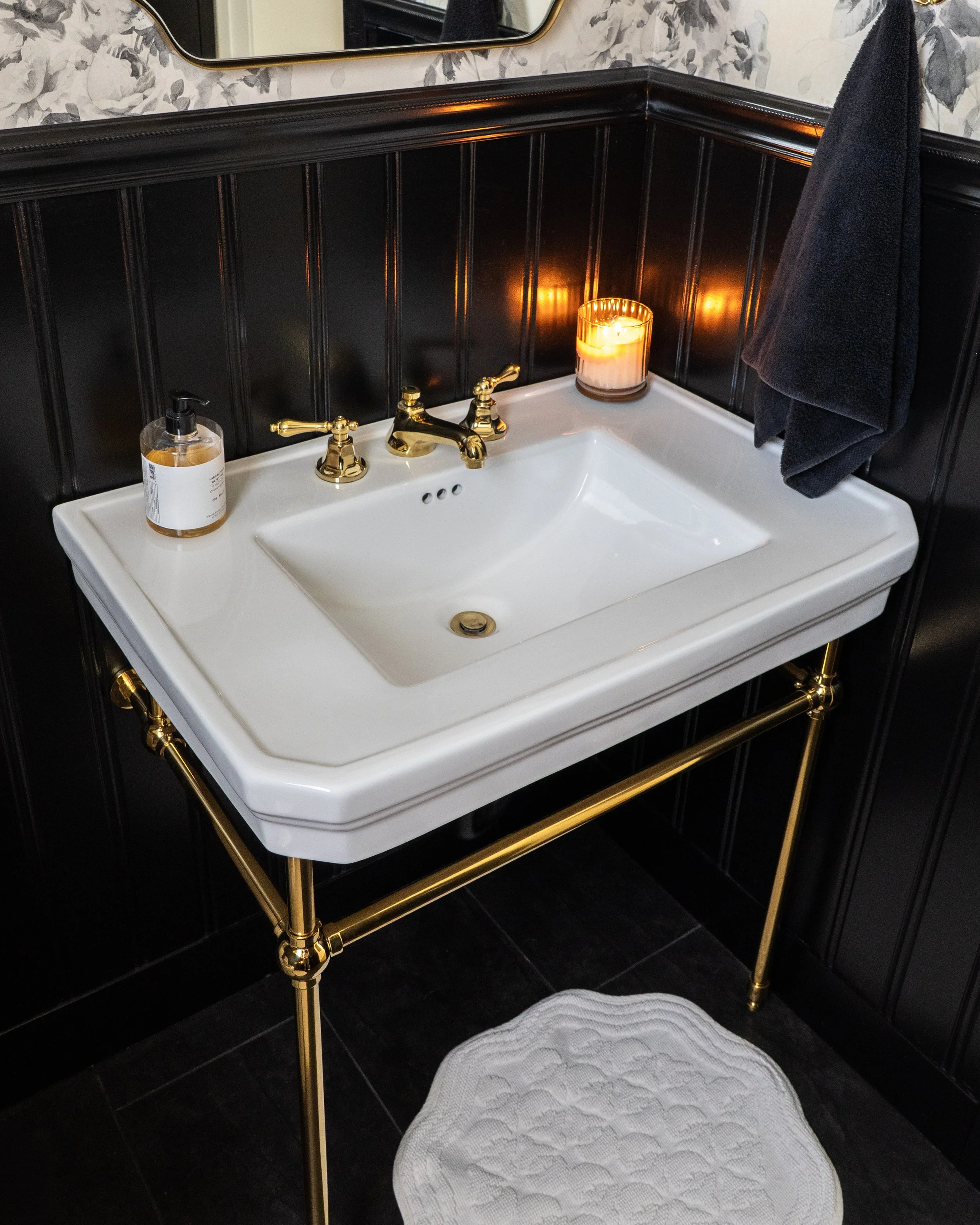 A bathroom sink with a black and gold vanity, a white ceramic basin, a tan candle burning, a black towel hanging on the wall, a bottle of liquid soap, a mirror above the sink, and a white textured rug on the dark flooring.