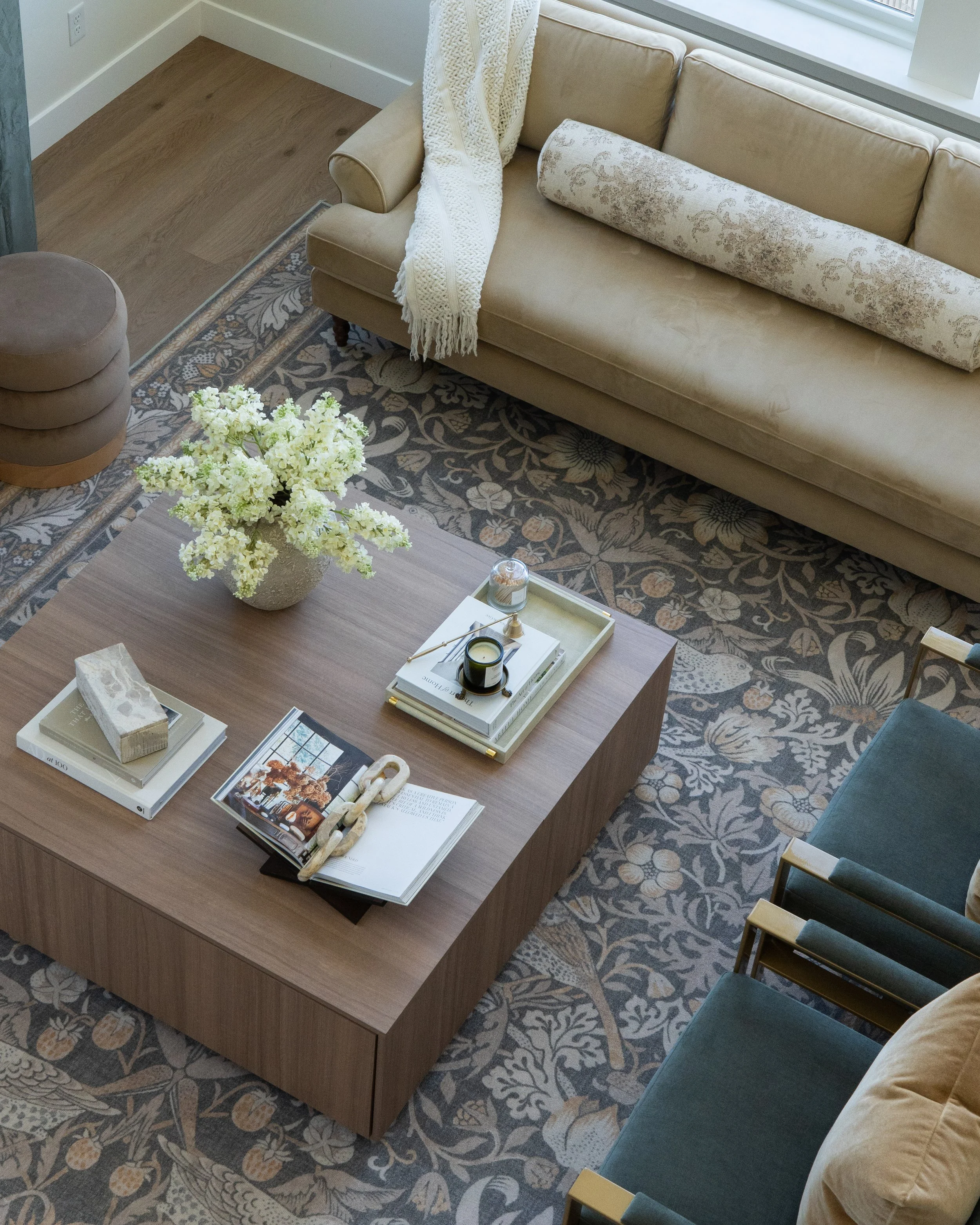 Living room with a wooden coffee table, beige sofa with decorative pillows and a blanket, a patterned area rug, a side chair with blue cushions, a vase with white flowers, books, and a window letting in natural light.
