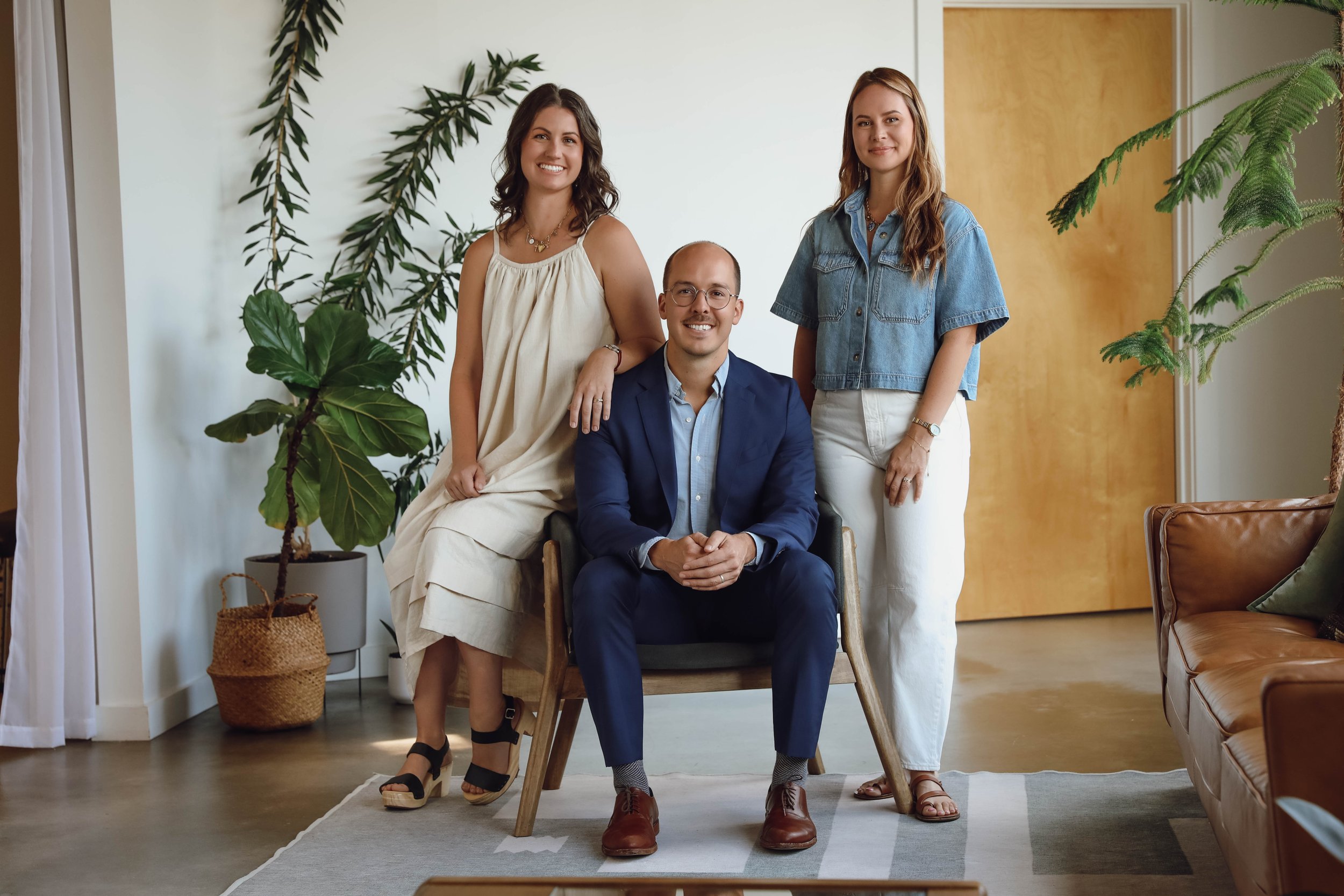 Three women and one man are inside a contemporary living room surrounded by indoor plants. The man, wearing a blue suit, is sitting in a chair with his hands clasped. The women, dressed casually, are standing and sitting near him, smiling at the camera.