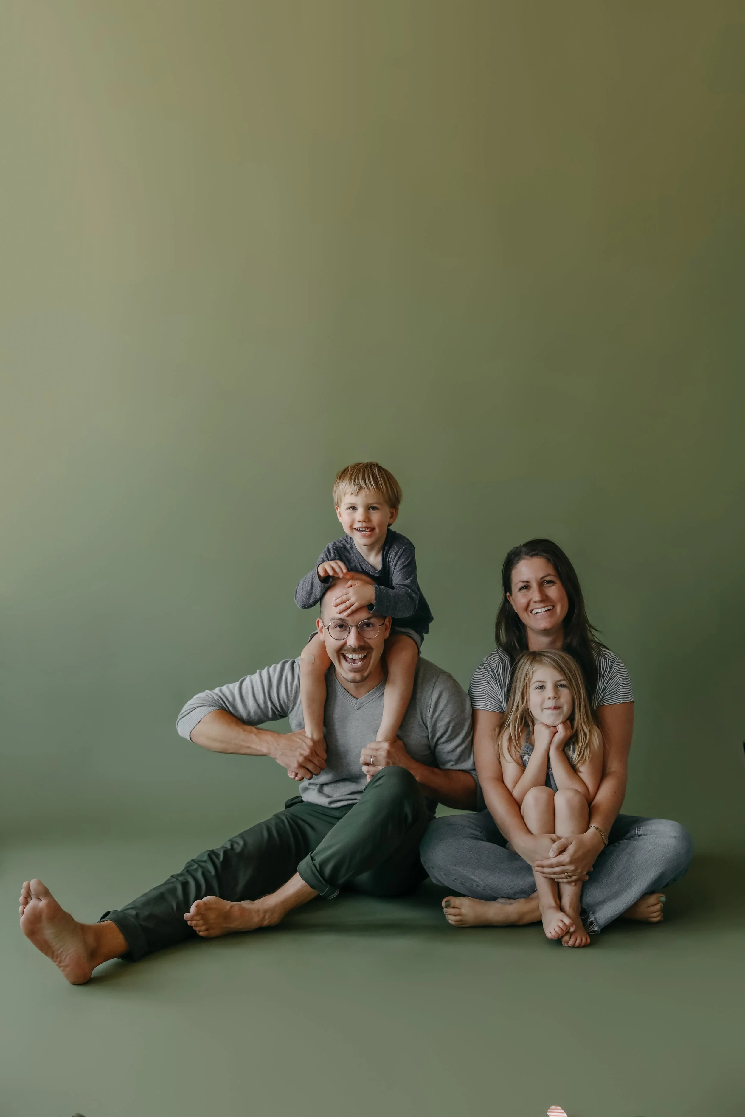 A family of five sitting and lying on the floor in front of a plain green backdrop, with a man carrying a young boy on his shoulders, and a woman and a girl sitting beside them, all smiling.
