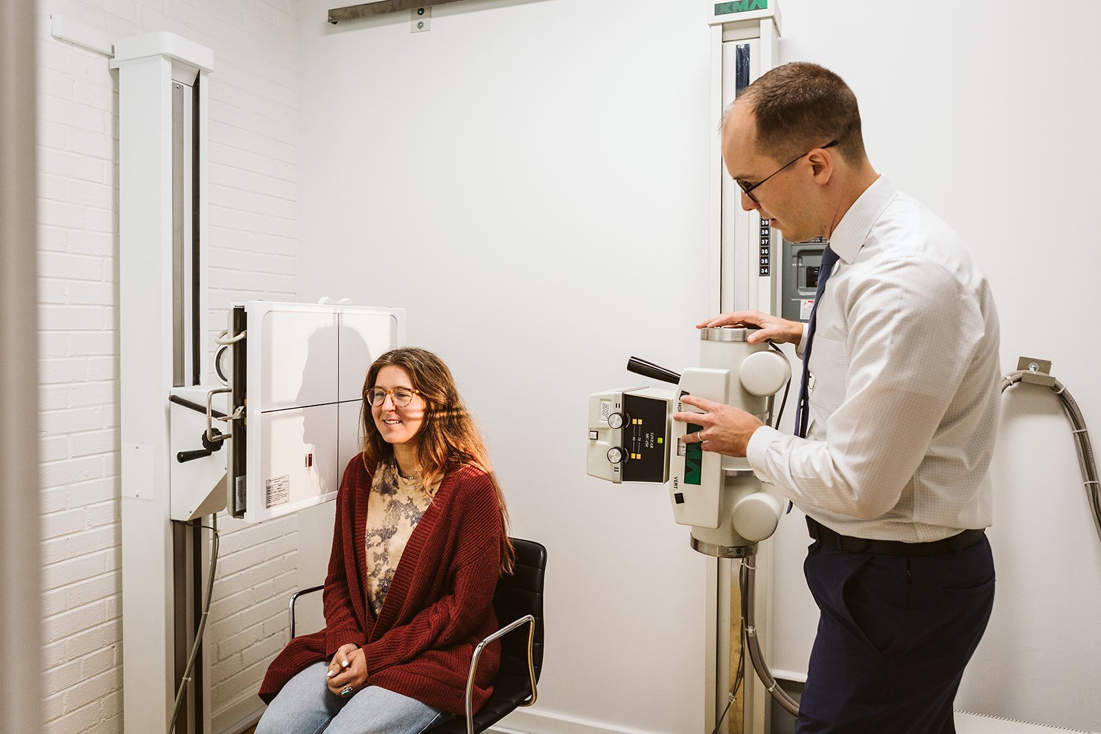 A woman in a maroon sweater is sitting in a chair in a medical office while a doctor in a white shirt and glasses uses a medical device on her head. The woman is smiling and looks relaxed. The room has white walls and medical equipment.