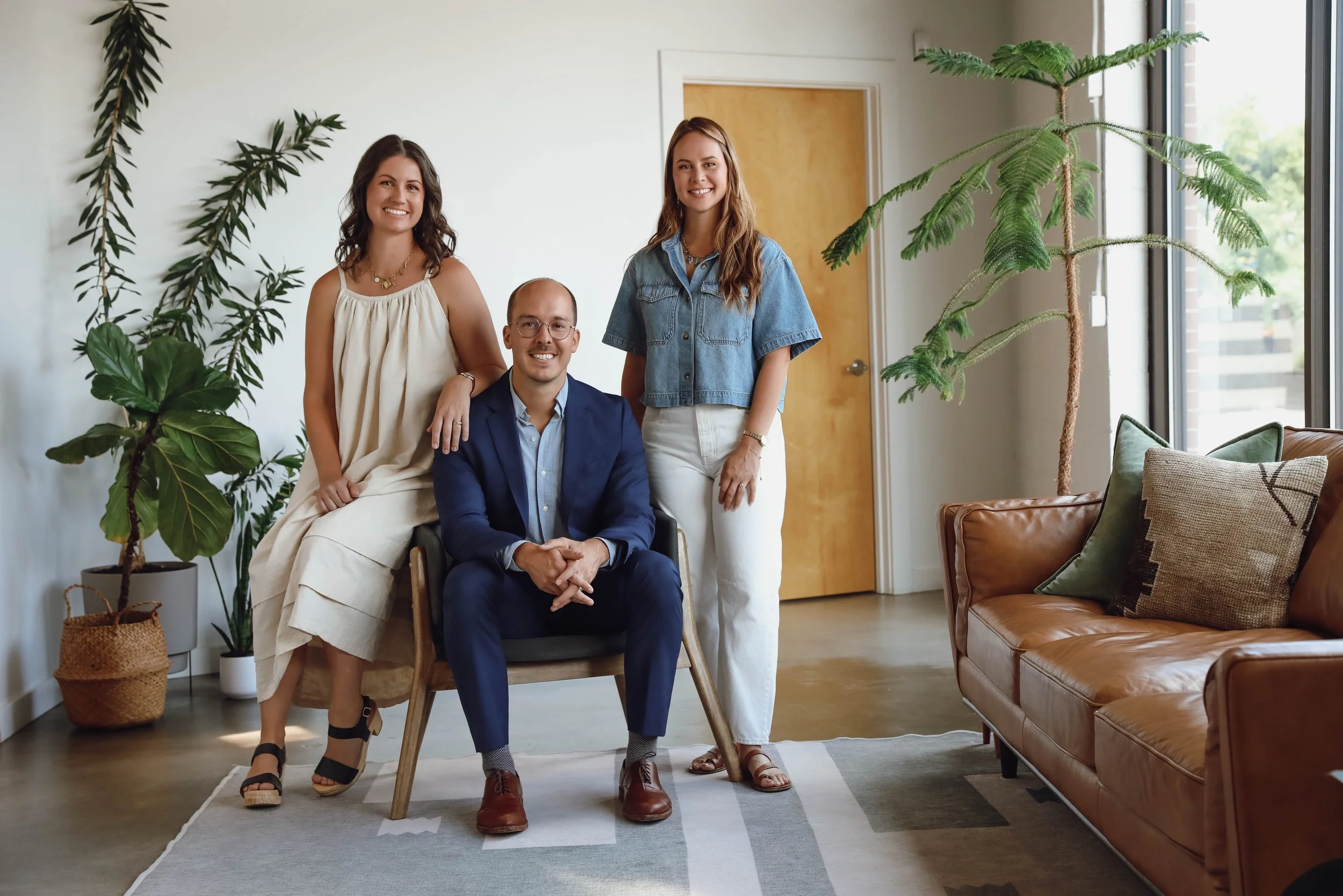 Three young adults in a bright, modern living room with large windows, surrounded by green plants, smiling and posing for the photo. One woman is sitting, and a man and a woman are standing beside and behind her.