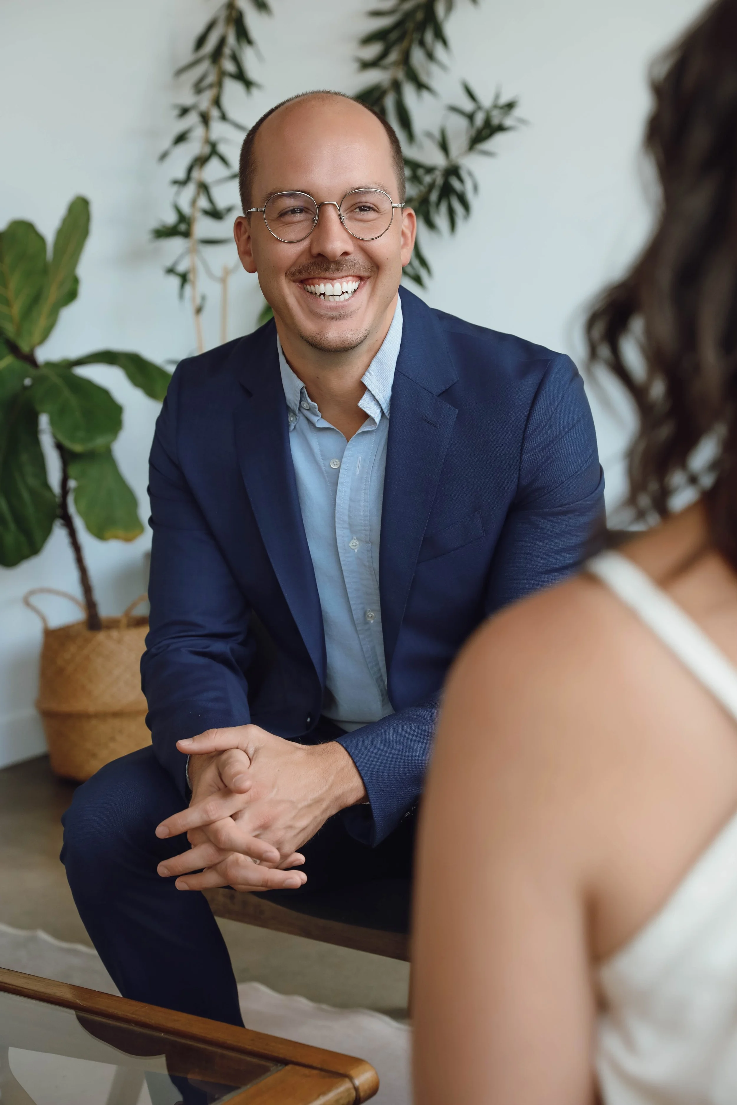 A man with glasses, a bald head, and a man bun, smiling and wearing a navy blazer and light shirt, engaged in conversation with a woman wearing a white tank top. They are sitting on a wooden bench in a room with plants in the background.