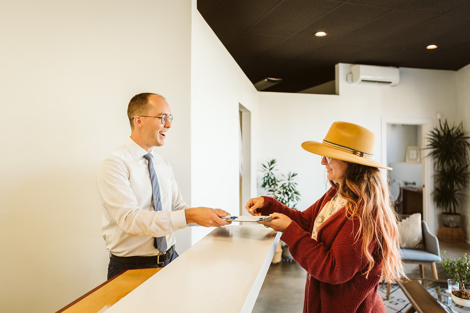 A man in a white shirt and tie at a reception desk handing a clipboard to a woman wearing a large tan hat and glasses, inside a modern, brightly lit room with plants and furniture.