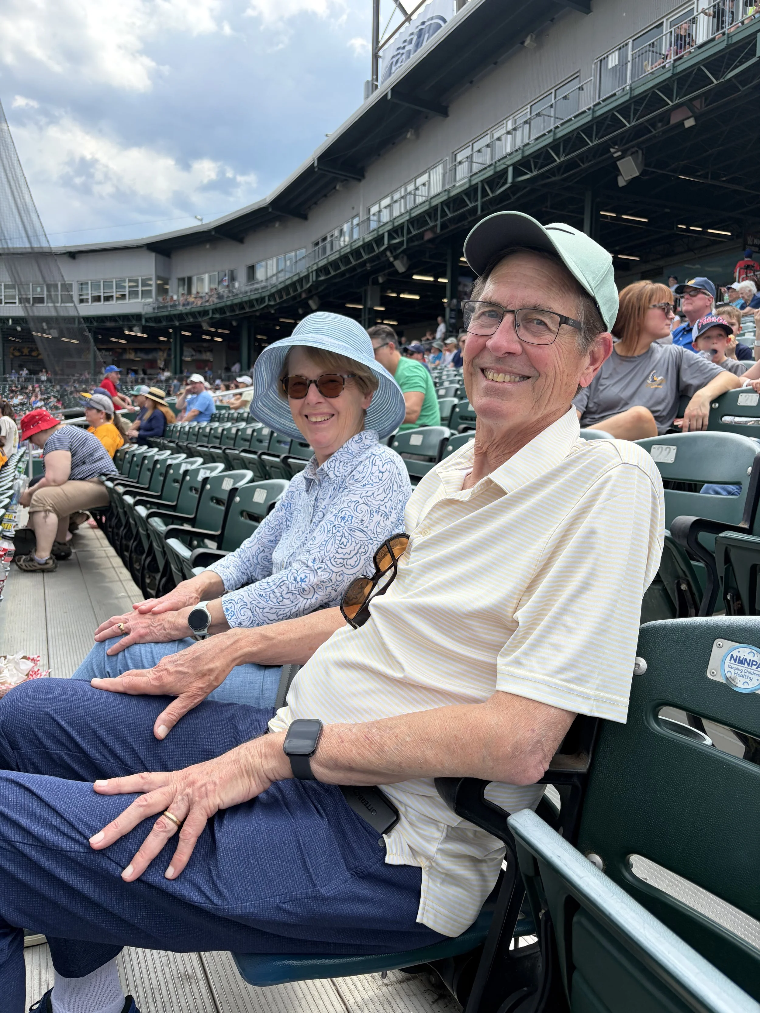 Two smiling elderly people, a woman wearing a light blue hat and sunglasses, and a man wearing a gray cap and glasses, sitting in a stadium with many other spectators around them, watching a sports event during daytime.