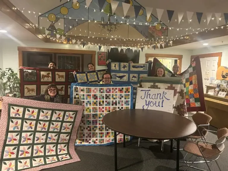 Group of people holding up various colorful quilts in a decorated room with string lights and bunting, with a round table in the foreground and a thank you sign visible.