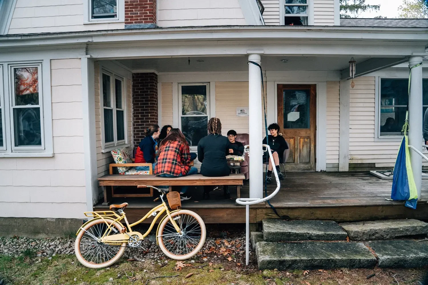 A group of people sitting on a porch outside a house, gathered around a small table, with a yellow bicycle parked in front and a colorful hammock hanging on the side.