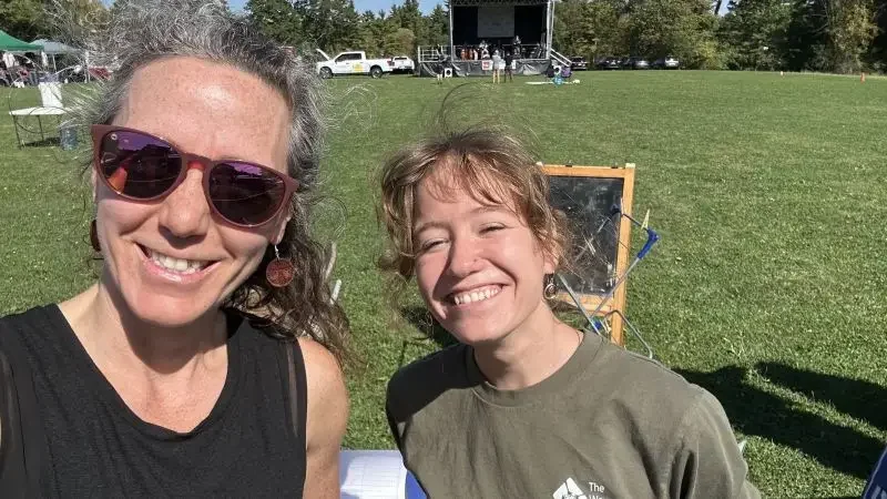 Two smiling women taking a selfie outdoors in a grassy field with a stage, vehicles, and trees in the background.