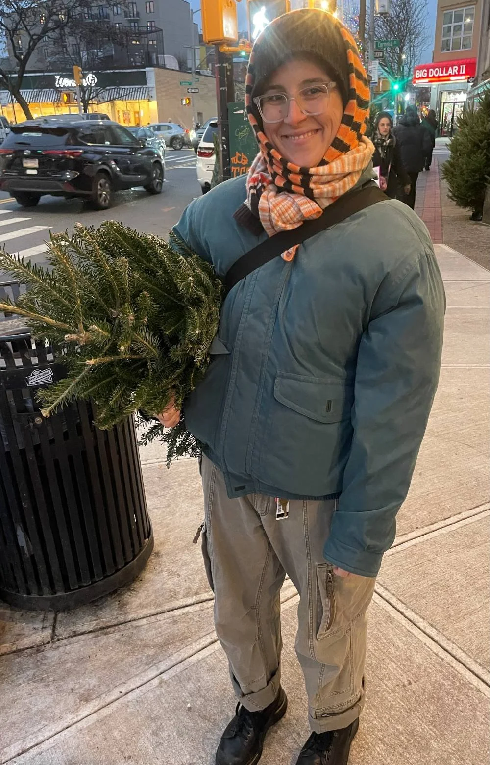 Person holding a small Christmas tree on a city sidewalk during evening, with cars and store signs in the background.