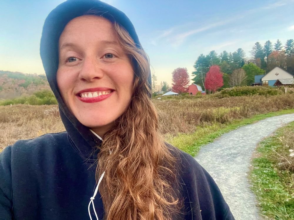 A young woman with long red hair wearing a black hoodie and a blue helmet, smiling while taking a selfie outdoors on a dirt path with trees and houses in the background during fall.