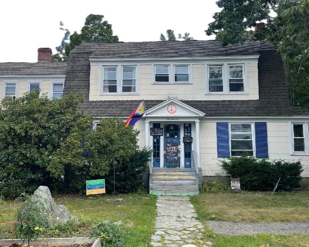 A two-story home with white exterior and blue shutters. A rainbow flag and a peace symbol are displayed near the front door. The entrance stairs lead to a porch with various signs, including one saying 'The People's Center'. There are bushes and a large rock in the front yard, with signs supporting social justice causes in the yard.