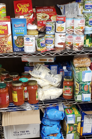 Shelf with canned soup, boxed mashed potatoes, chicken broth, and other canned goods, jars of pasta sauce, and grocery items in bags.