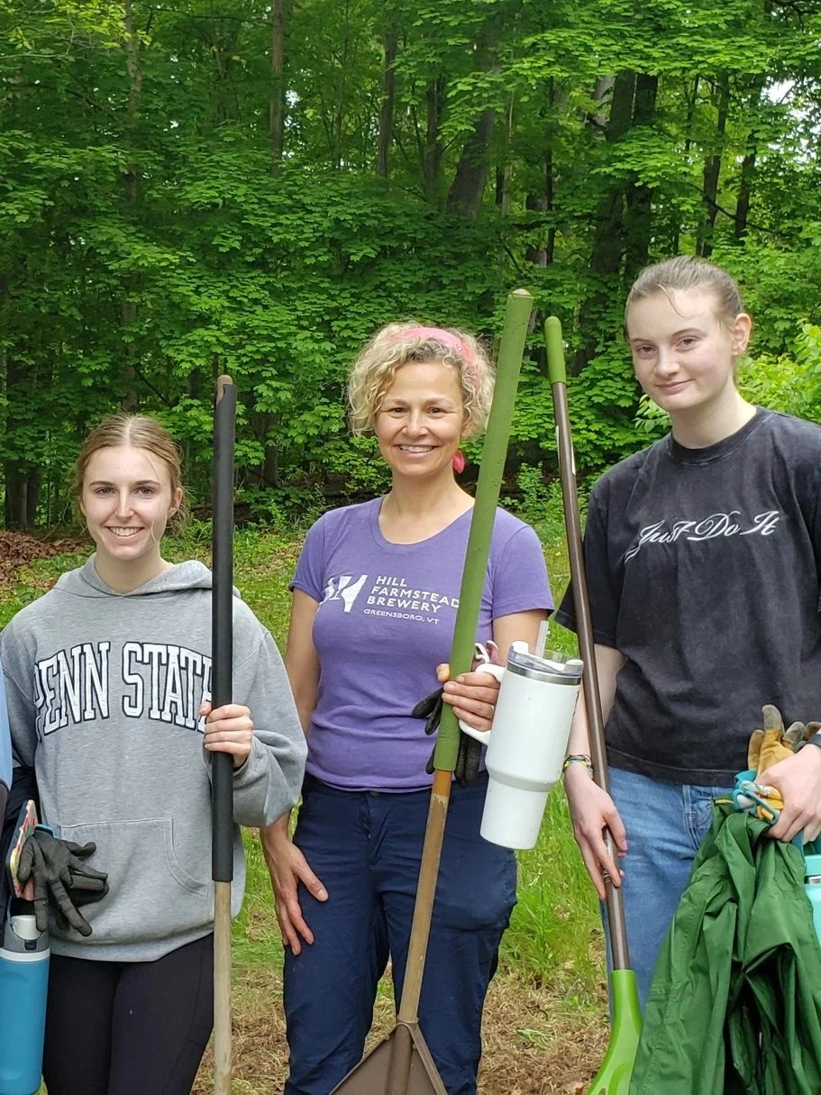 Three people standing outdoors in a lush green forest, holding gardening tools and smiling at the camera.