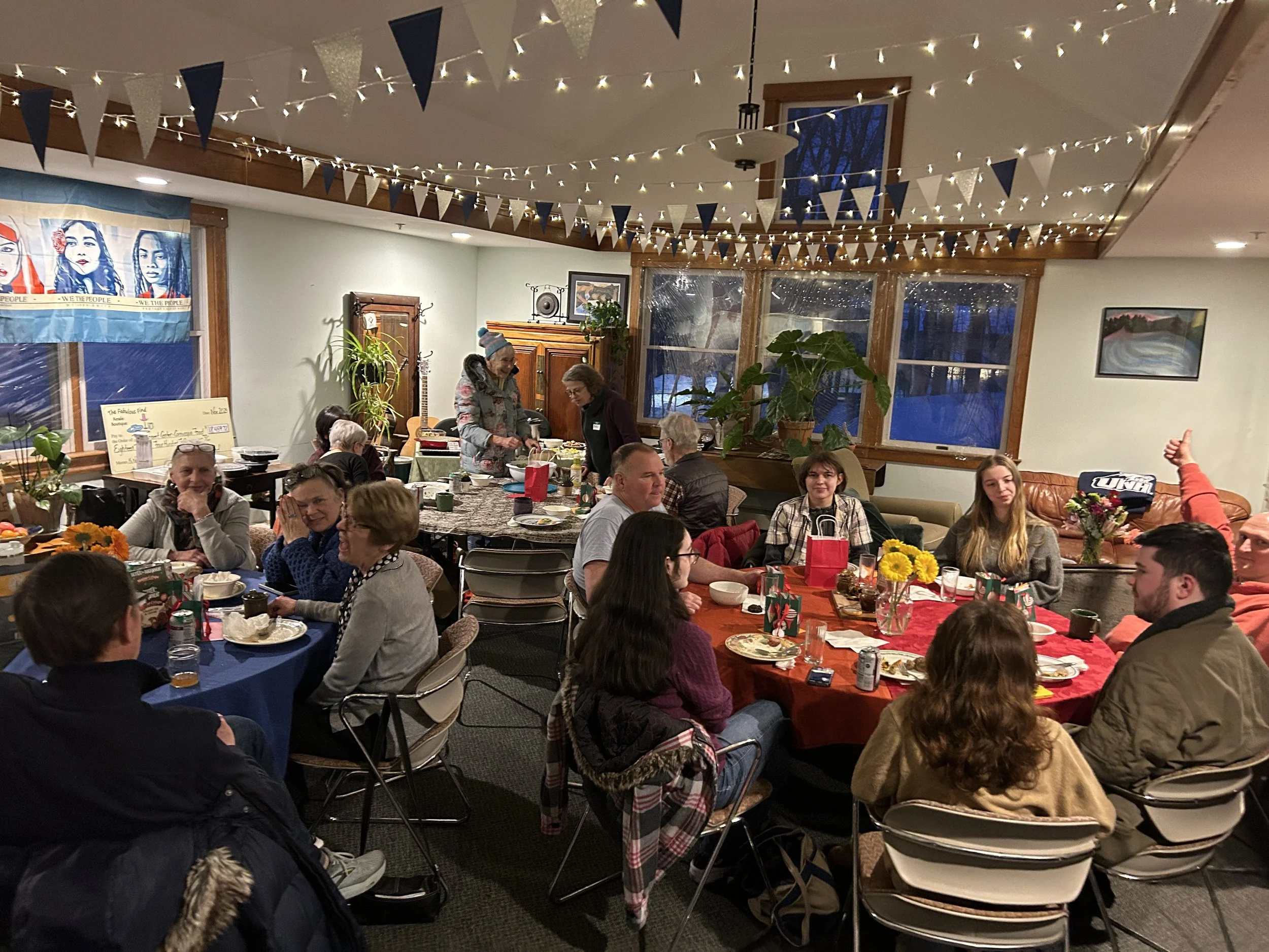 Indoor gathering with people seated at tables decorated with flowers, serving food and drinks, under string lights and festive bunting, with large windows showing a winter scene outside.