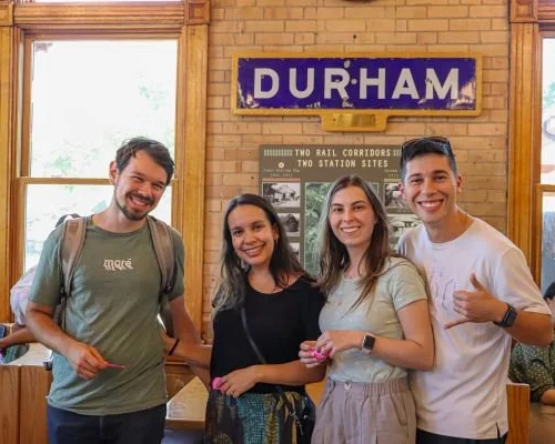 Four smiling young adults standing inside a train station near a sign that reads 'DURHAM.' They are posing for a photo, with the group consisting of two men and two women. One man is giving a thumbs-up gesture.