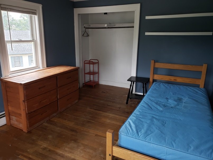 A simple room with blue walls, a window, a wooden dresser, an empty closet with a red rolling cart, a small black side table, and a bed with a blue mattress.