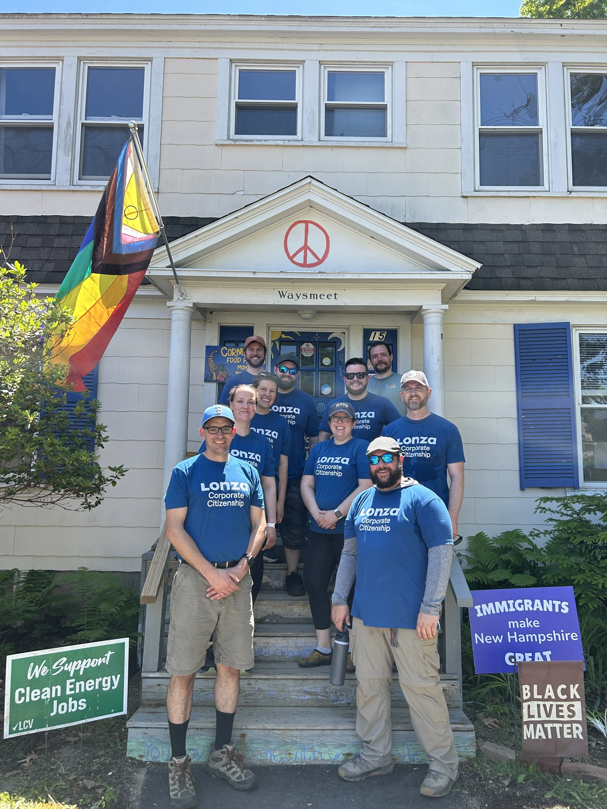Group of people standing on front steps of a building with a peace symbol and window above, surrounded by signs with messages about supporting clean energy jobs, immigrant rights, and Black Lives Matter.