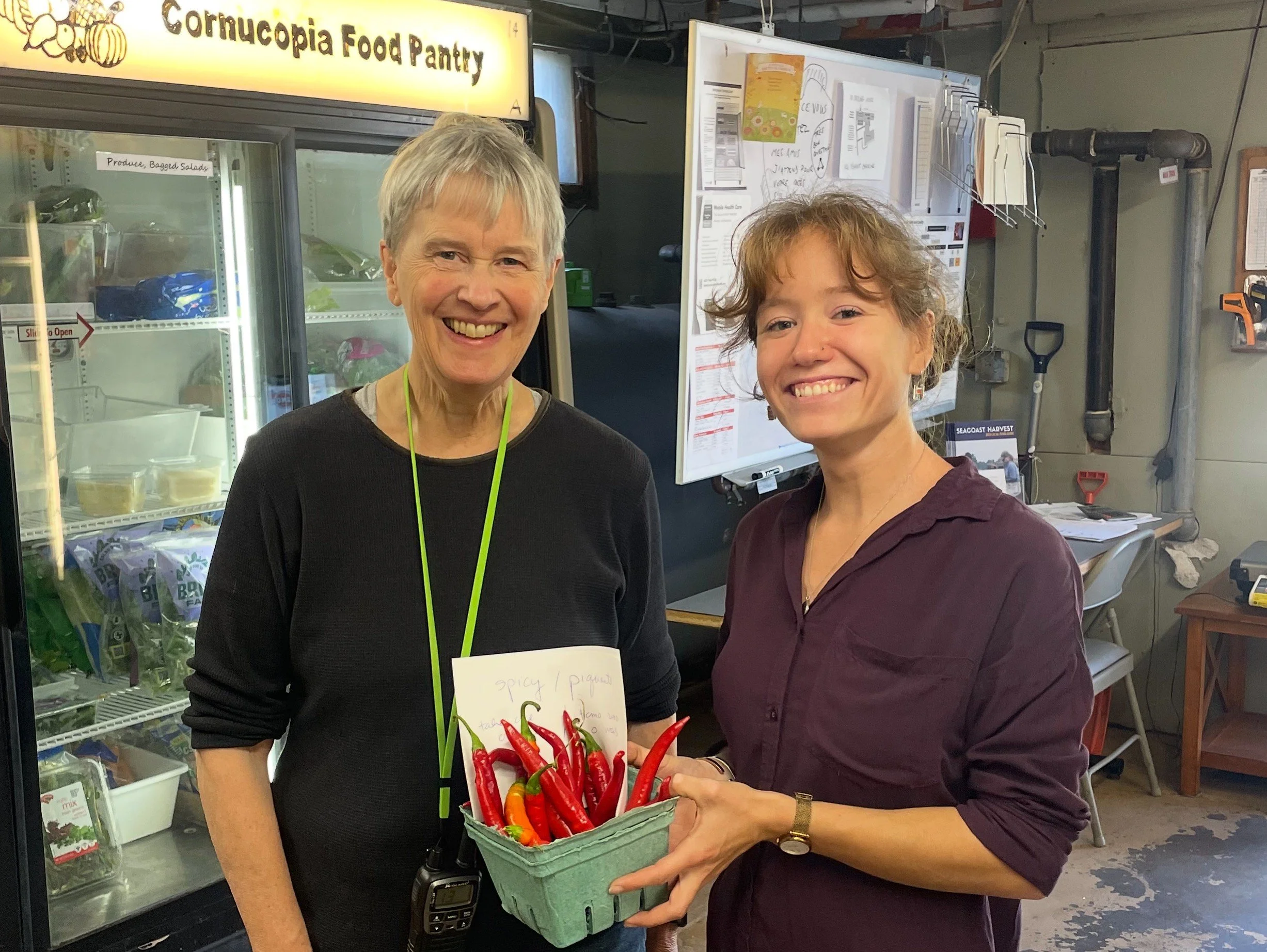 Two women smiling and holding a basket of red chili peppers at the Cornucopia Food Pantry.
