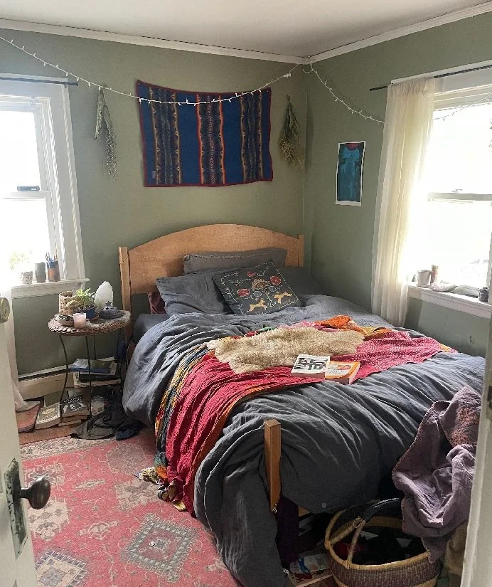 A cozy bedroom with green walls, a wooden headboard, and a bed covered with colorful blankets and pillows. Two windows with white curtains let in natural light. A small round side table holds various items, and a pink patterned rug covers the floor. Decor includes string lights and wall art.