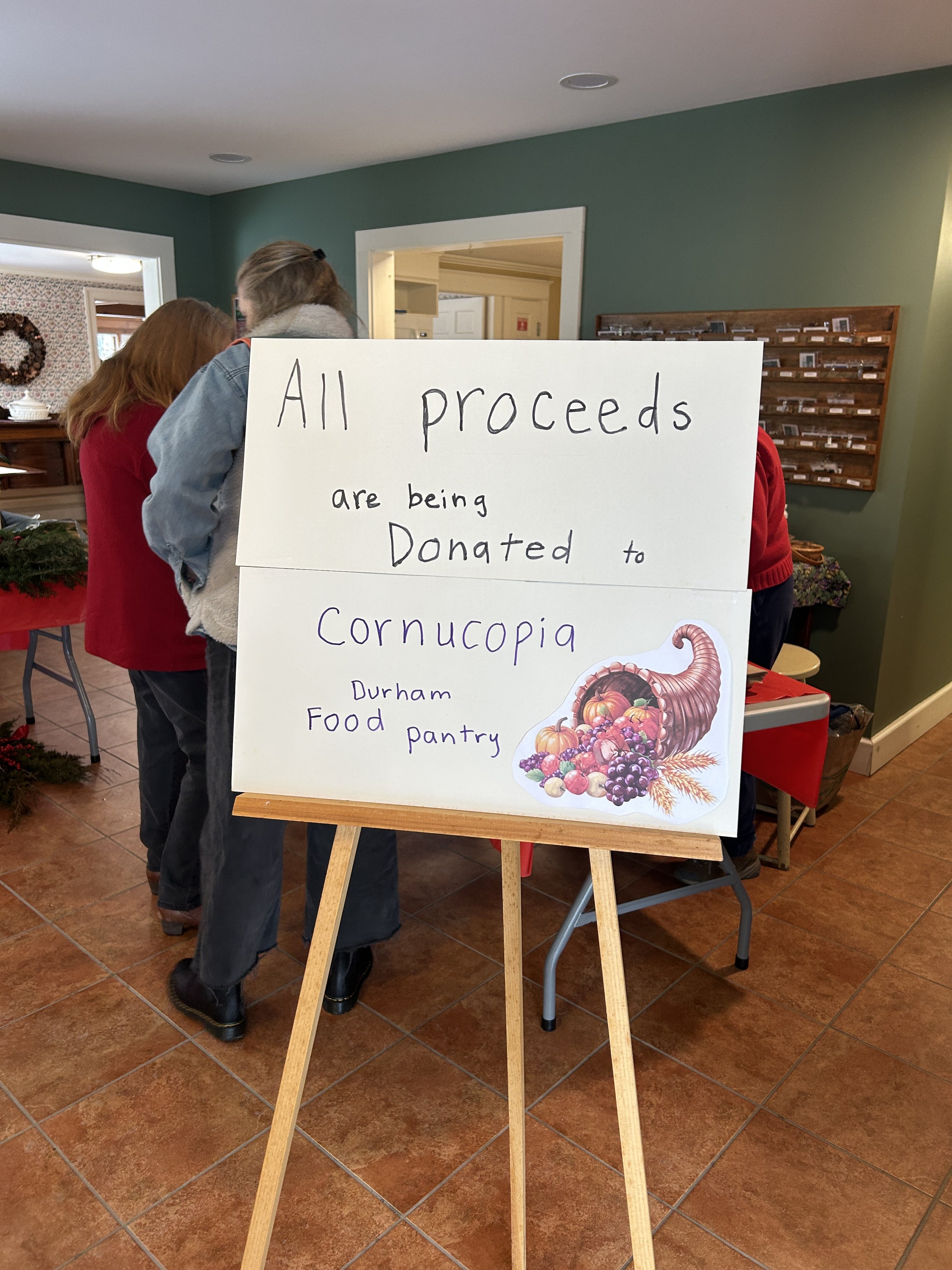A large sign that reads 'All proceeds are being donated to Cornucopia Durham Food Pantry' with an illustration of a cornucopia filled with fruits and vegetables. The setting is indoors at the Durham Unitarian Universalist Fellowship.