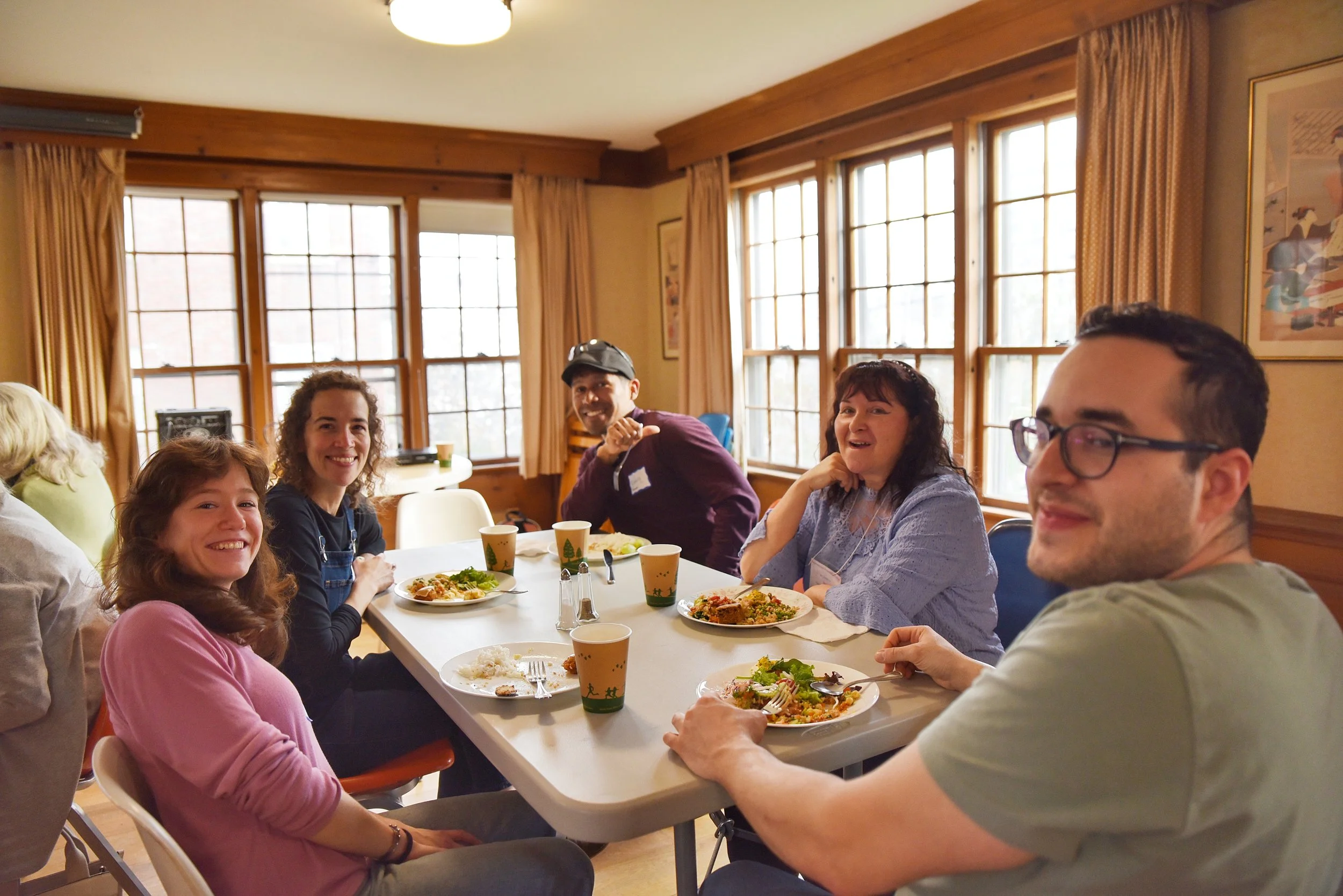 Group of five people sitting around a table, smiling, with plates of food and cups, in a room with large windows and wooden decor.