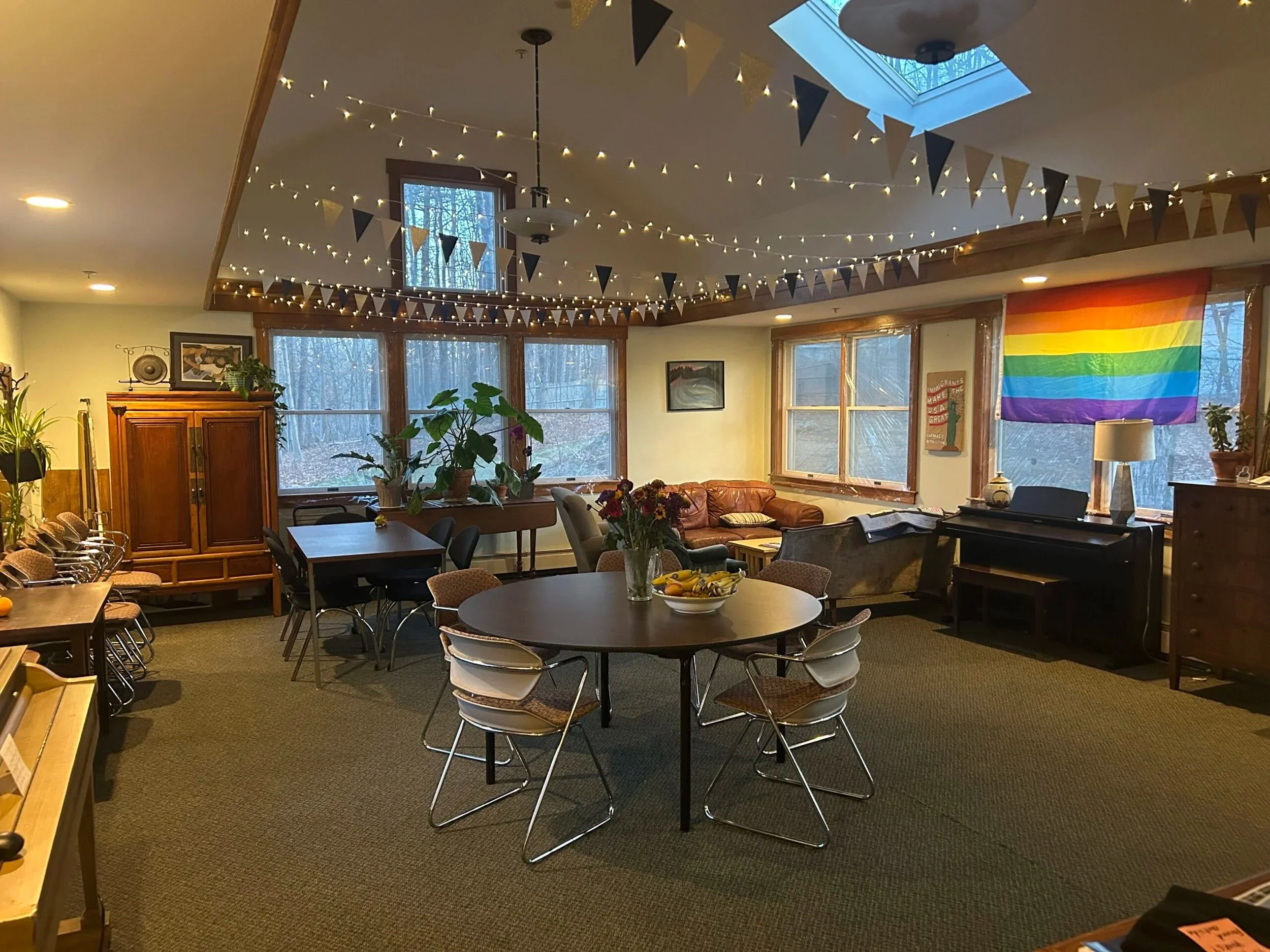 A decorated indoor space with string lights and black and tan pennant banners hanging from the ceiling. There are several large windows and a skylight letting in natural light. A table with a vase of flowers and bananas is in the foreground, and a colorful rainbow pride flag hangs on the wall to the right.