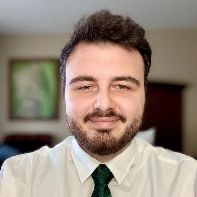 A man with dark hair, a beard, and mustache wearing a white shirt and a dark green tie, smiling in a room with a window and some blurred furnishings in the background.