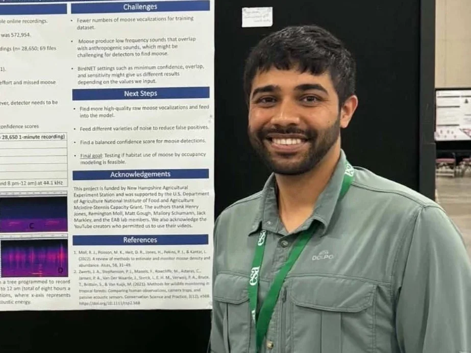 A man with short dark hair and a beard, smiling, wearing a gray button-up shirt with a green lanyard, standing next to a scientific poster presentation.