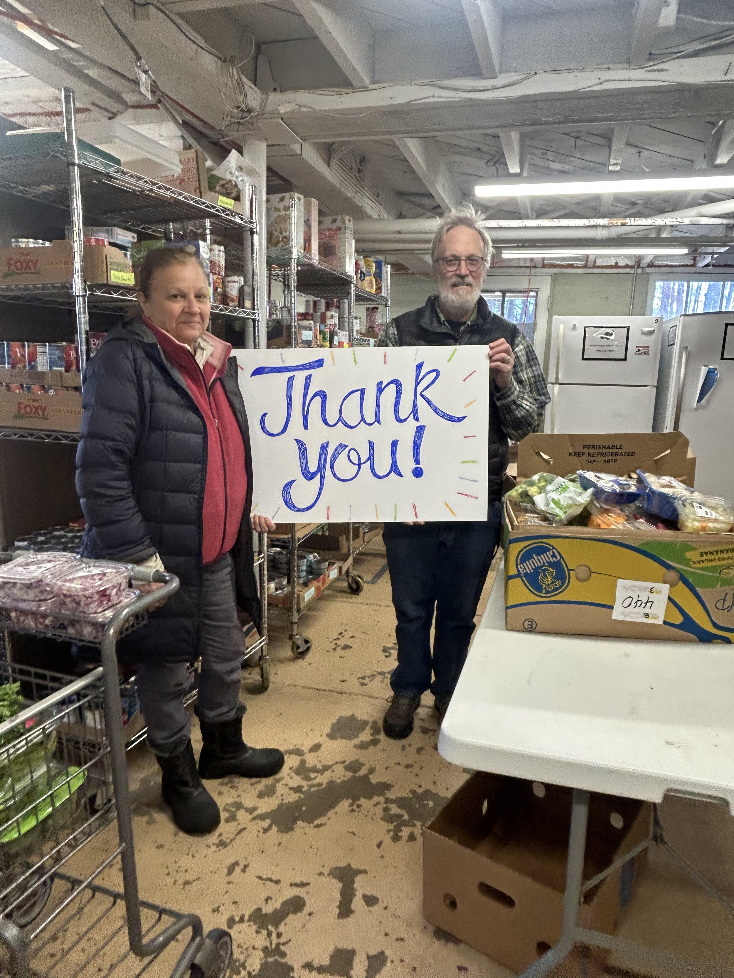 Two people are standing inside Cornucopia Food Pantry, holding a large white sign with blue handwritten text that says 'Thank You!'.