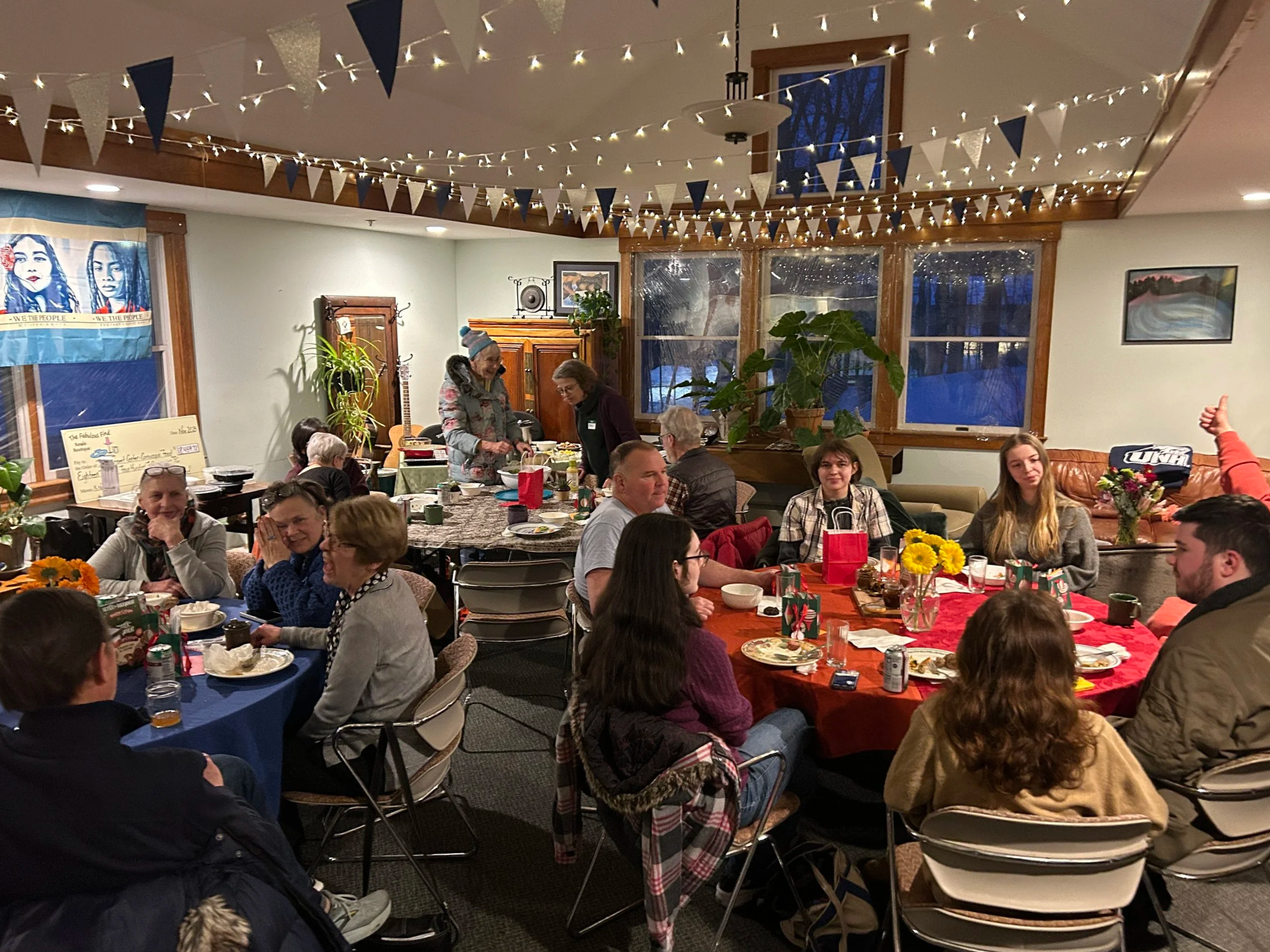 A lively indoor gathering with people seated at decorated tables, enjoying a festive meal. The room has string lights and blue and gold bunting hanging from the ceiling. There is a window with a view of a snowy outdoor scene, and various decorations and art on the walls.