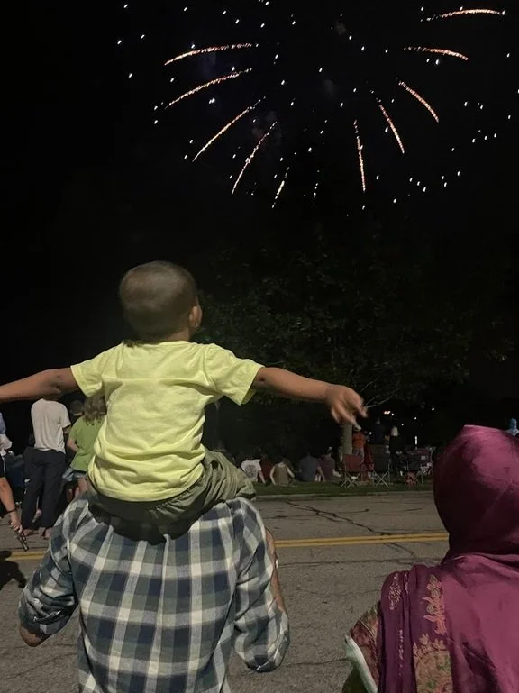 Child sitting on an adult's shoulders watching fireworks in the night sky during a celebration or event.