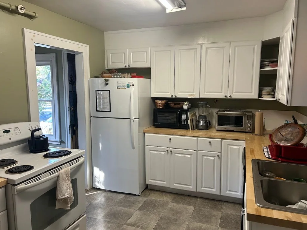 Kitchen with white cabinets, a white refrigerator, a stove, a microwave, a blender, a toaster oven, and a sink with dishes, with an open window showing trees outside.