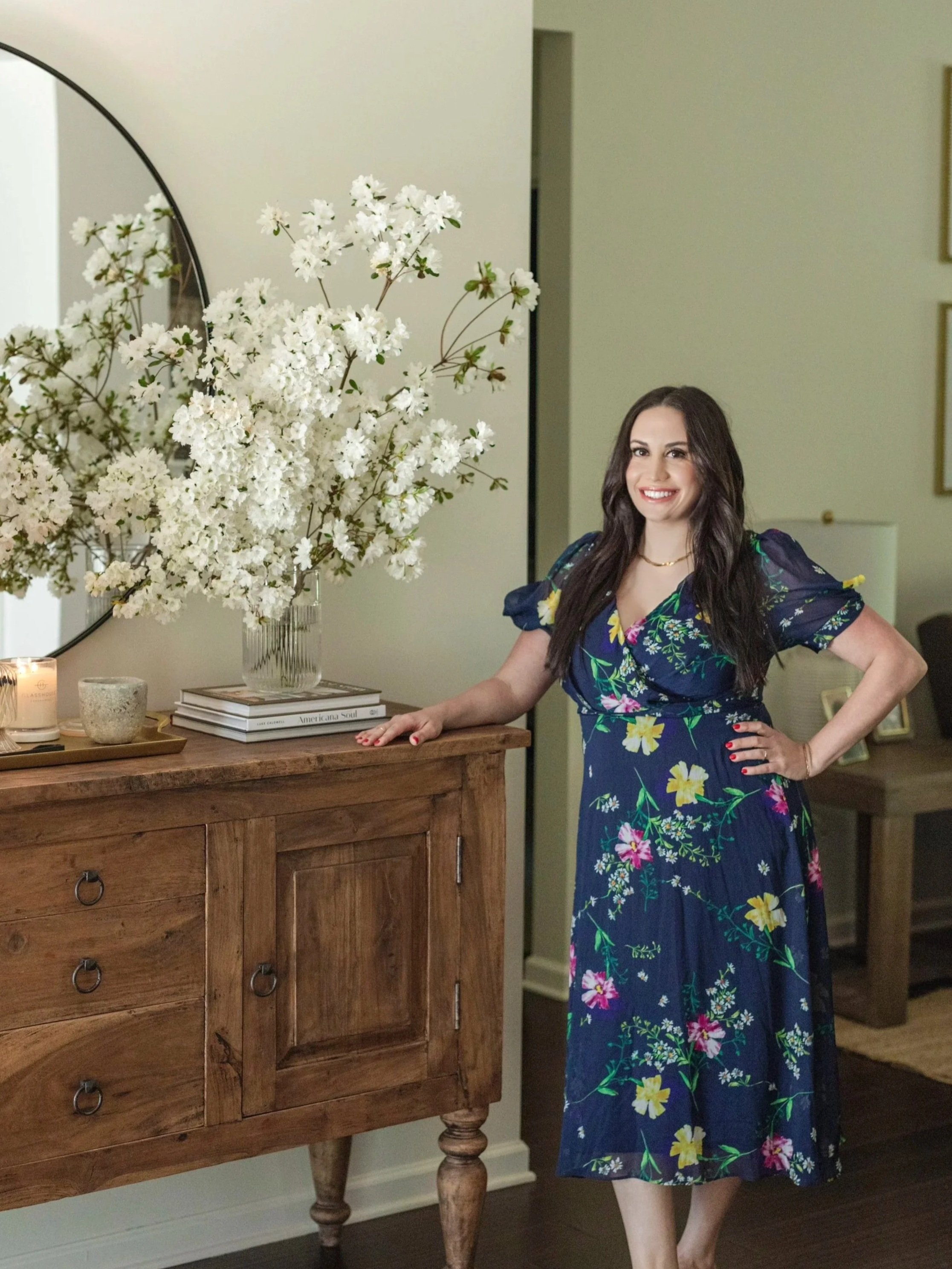 A woman with long dark hair and a bright smile, wearing a blue floral dress, posing in a room with a wooden dresser topped with flowers and books, and a mirror.