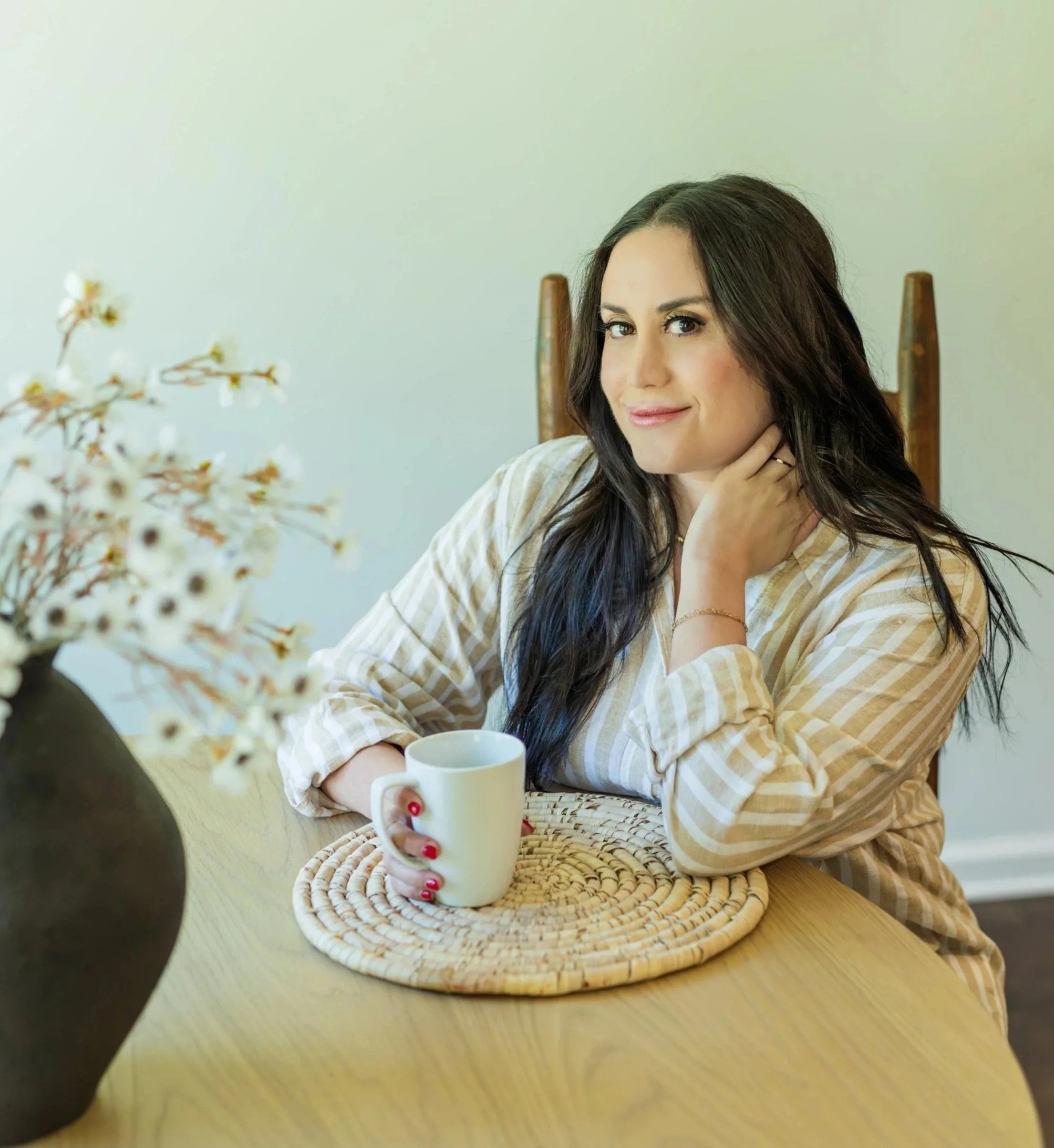 A woman with dark hair and light skin sitting at a wooden table, holding a white mug, with a blurred plant in the foreground and a plain light-colored wall in the background.
