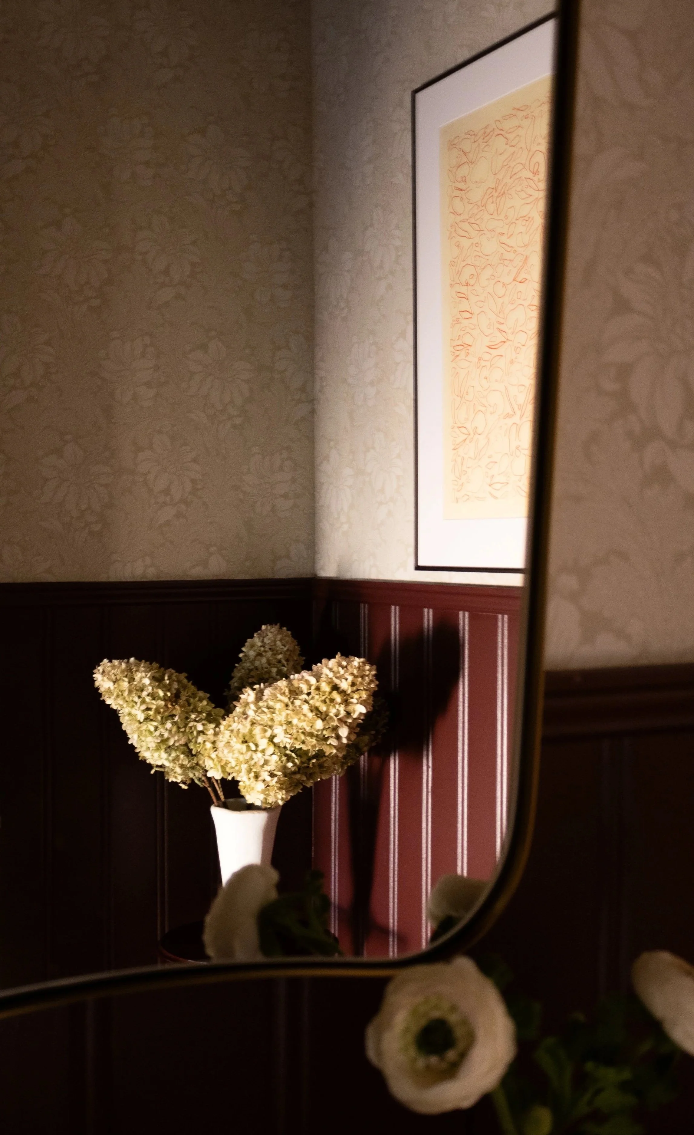 A mirror reflecting a wall with beige floral wallpaper and a framed abstract artwork with orange lines, a white vase with dried cream-colored flowers, and additional white flowers in the foreground.