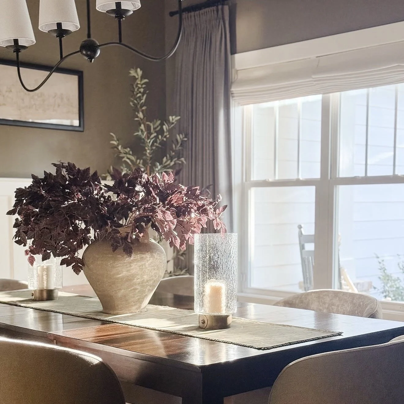 A dining room table with a large vase of dark purple leaves, a candle in a glass holder, and a table runner, with a large window in the background letting in sunlight.
