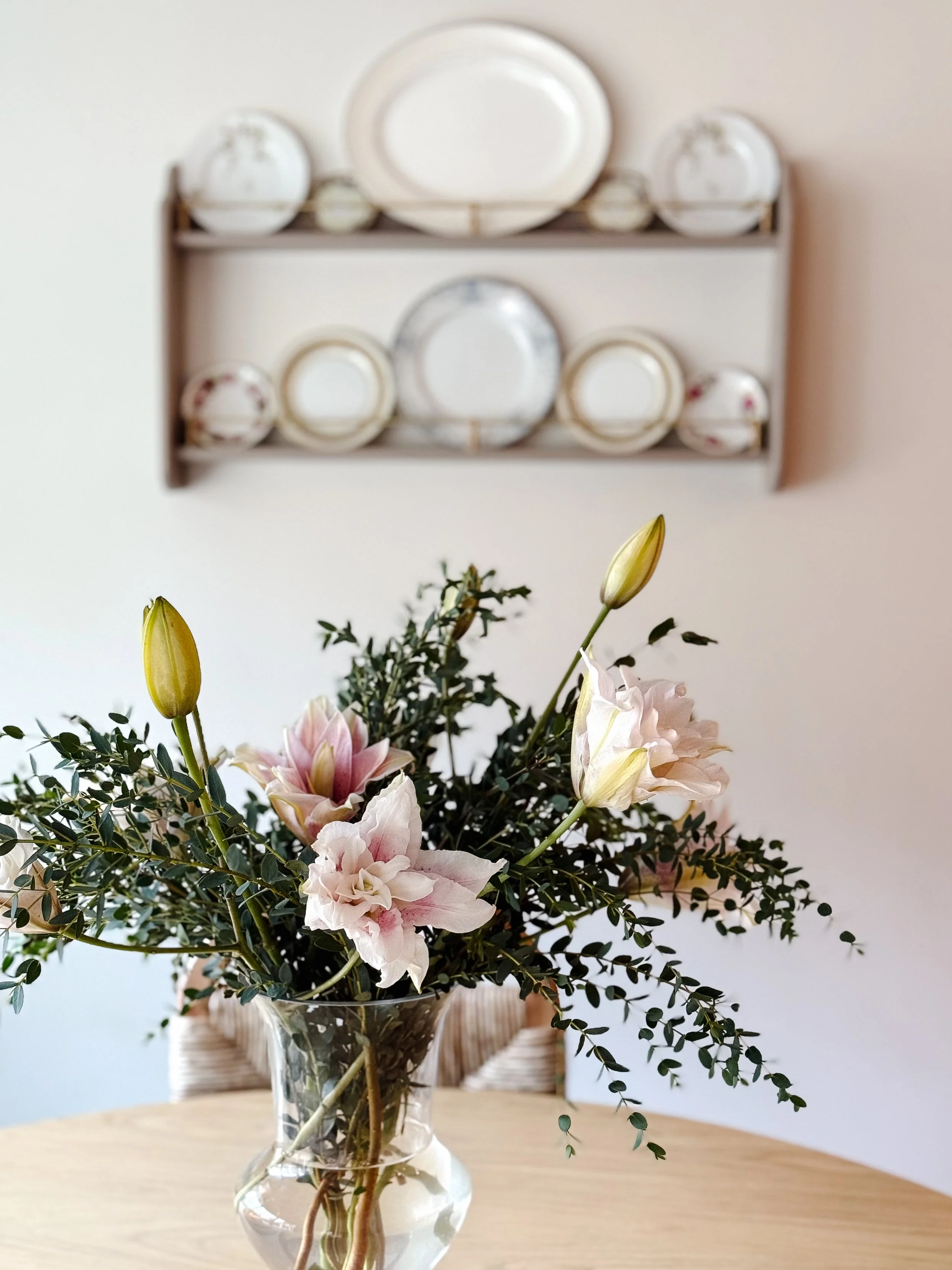 A glass vase with a bouquet of pink and white flowers and greenery on a wooden table, with a blurred wall and a cabinet with plates in the background.
