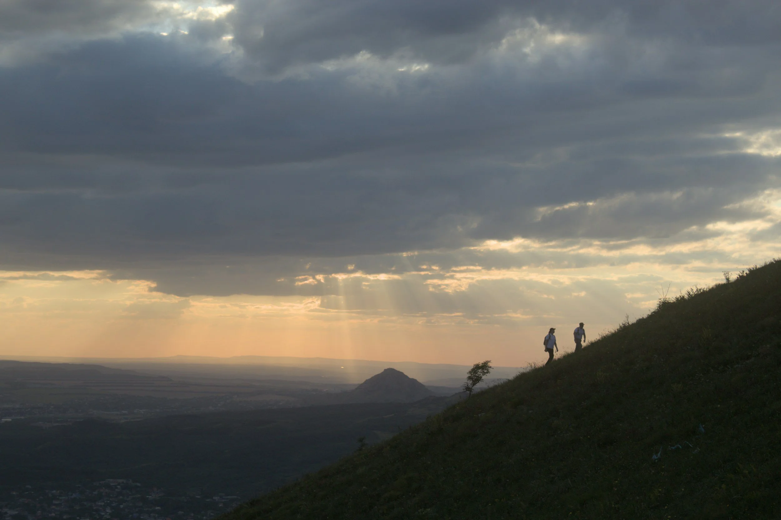 Two hikers walking along a hillside during sunset with a cloudy sky overhead.