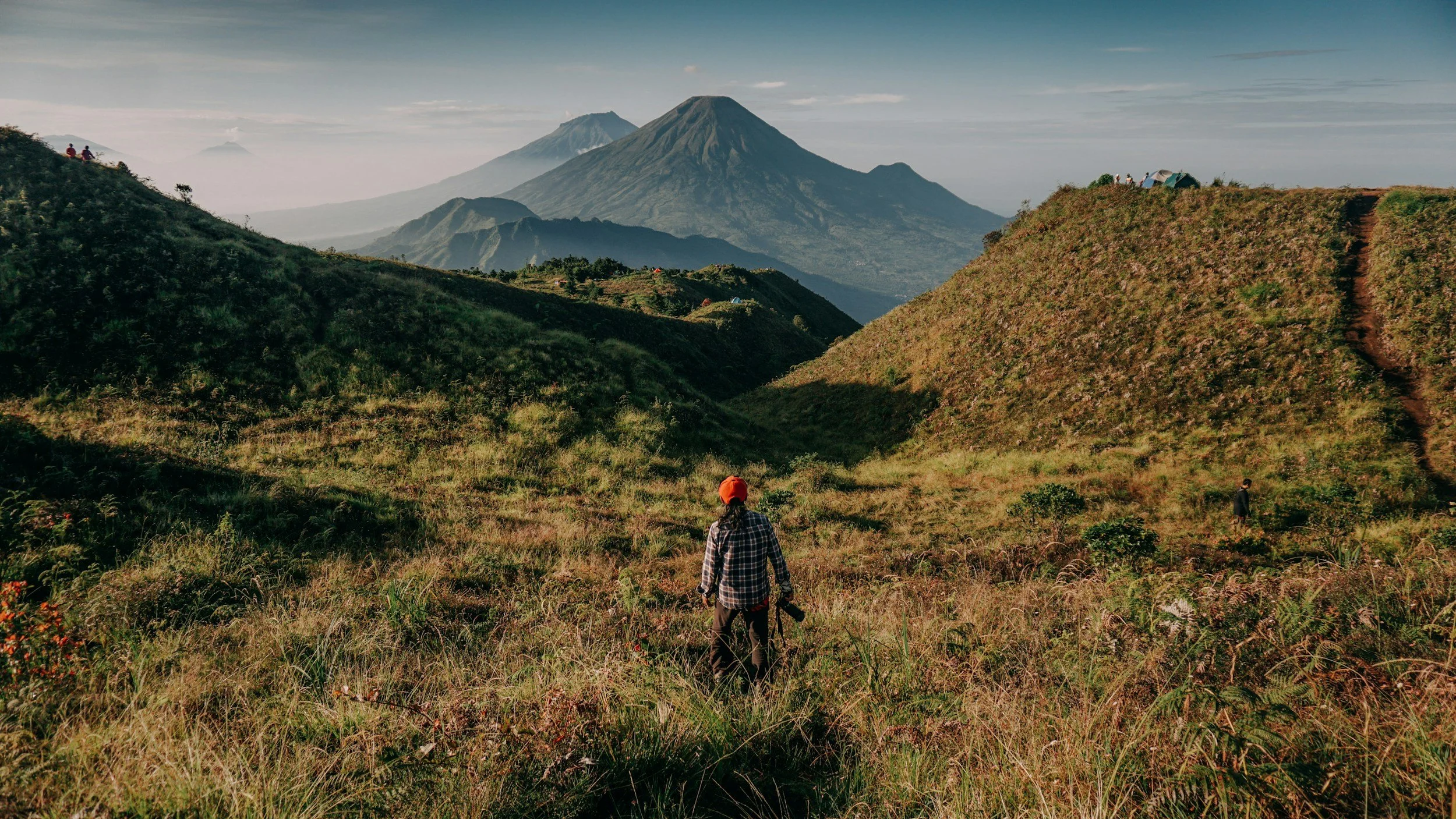 A person wearing a hat and checkered shirt carrying a camera on a hiking trail in a lush green mountainous area with volcanic mountains in the background.