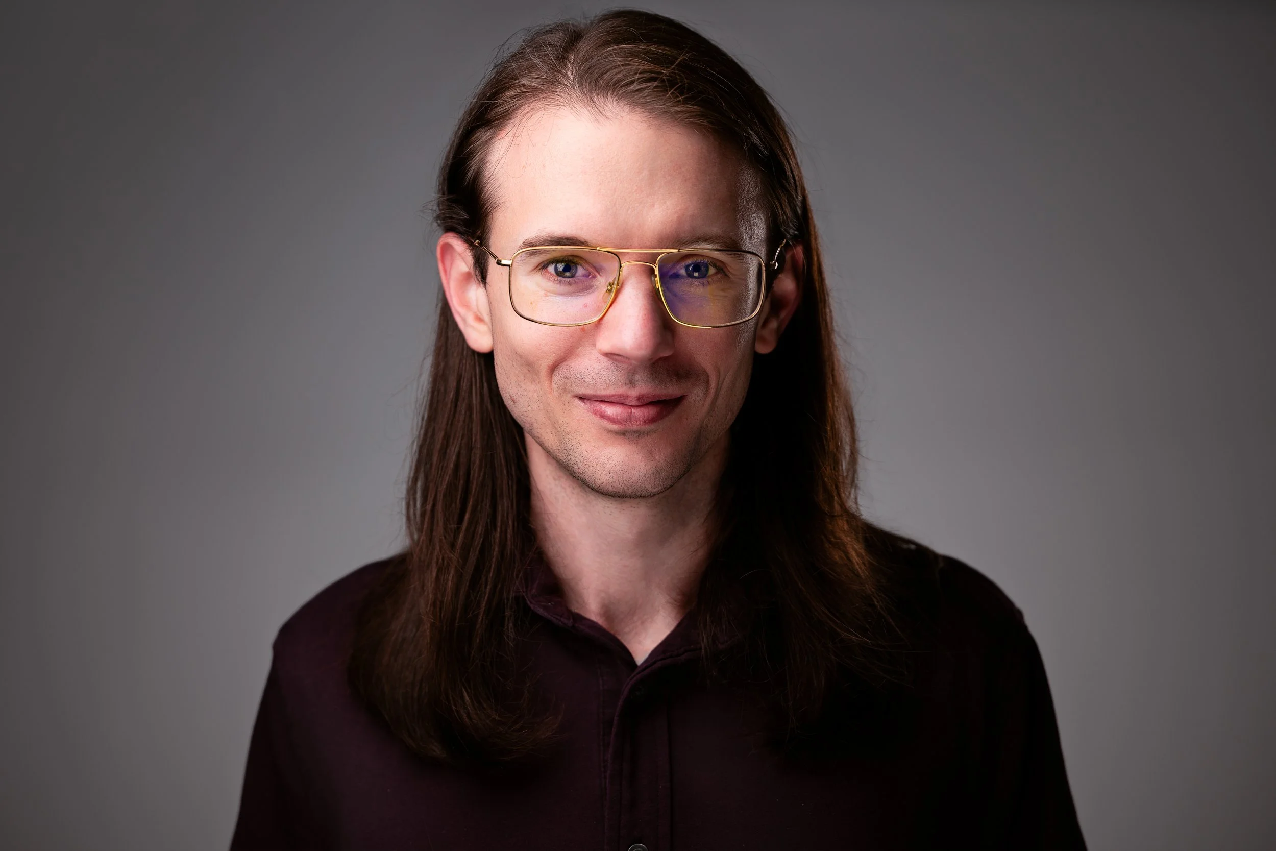 A man with long brown hair, wearing glasses and a dark-colored shirt, smiling slightly, against a plain gray background.