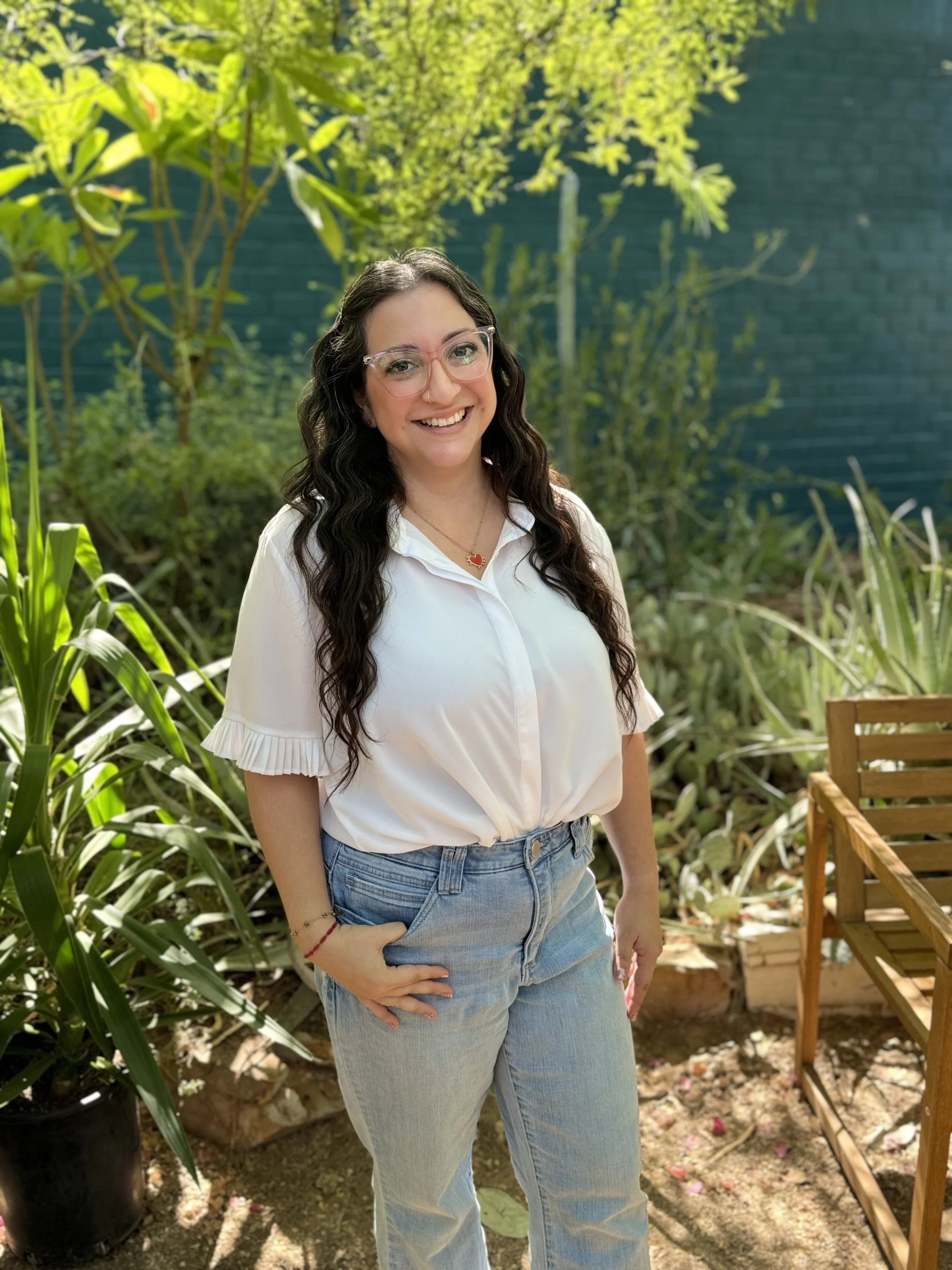 A woman with long dark wavy hair, wearing glasses, a white shirt, and blue jeans, smiling outdoors in a garden with green plants and a blue wall in the background.