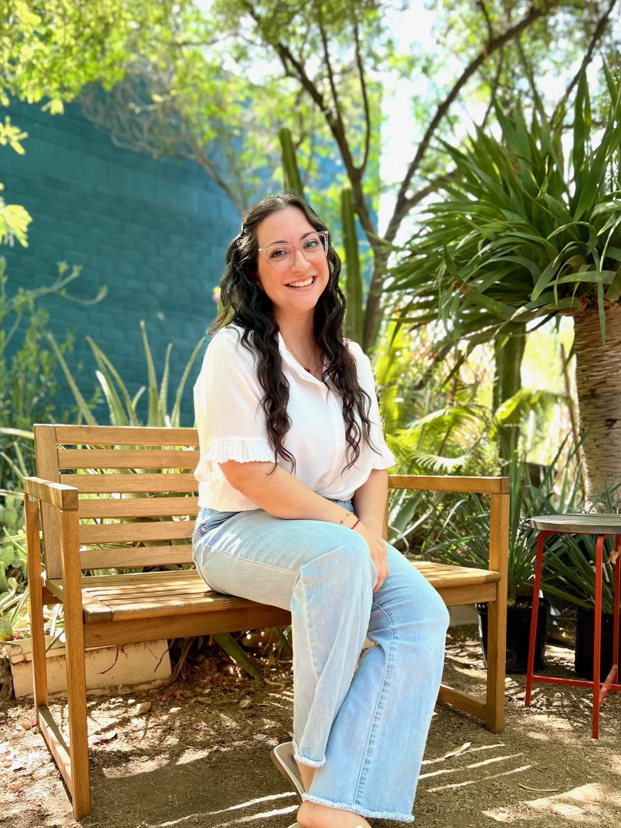 A woman with long dark curly hair, wearing glasses and a white blouse, sitting on a wooden bench outdoors surrounded by lush green plants and trees, smiling at the camera.