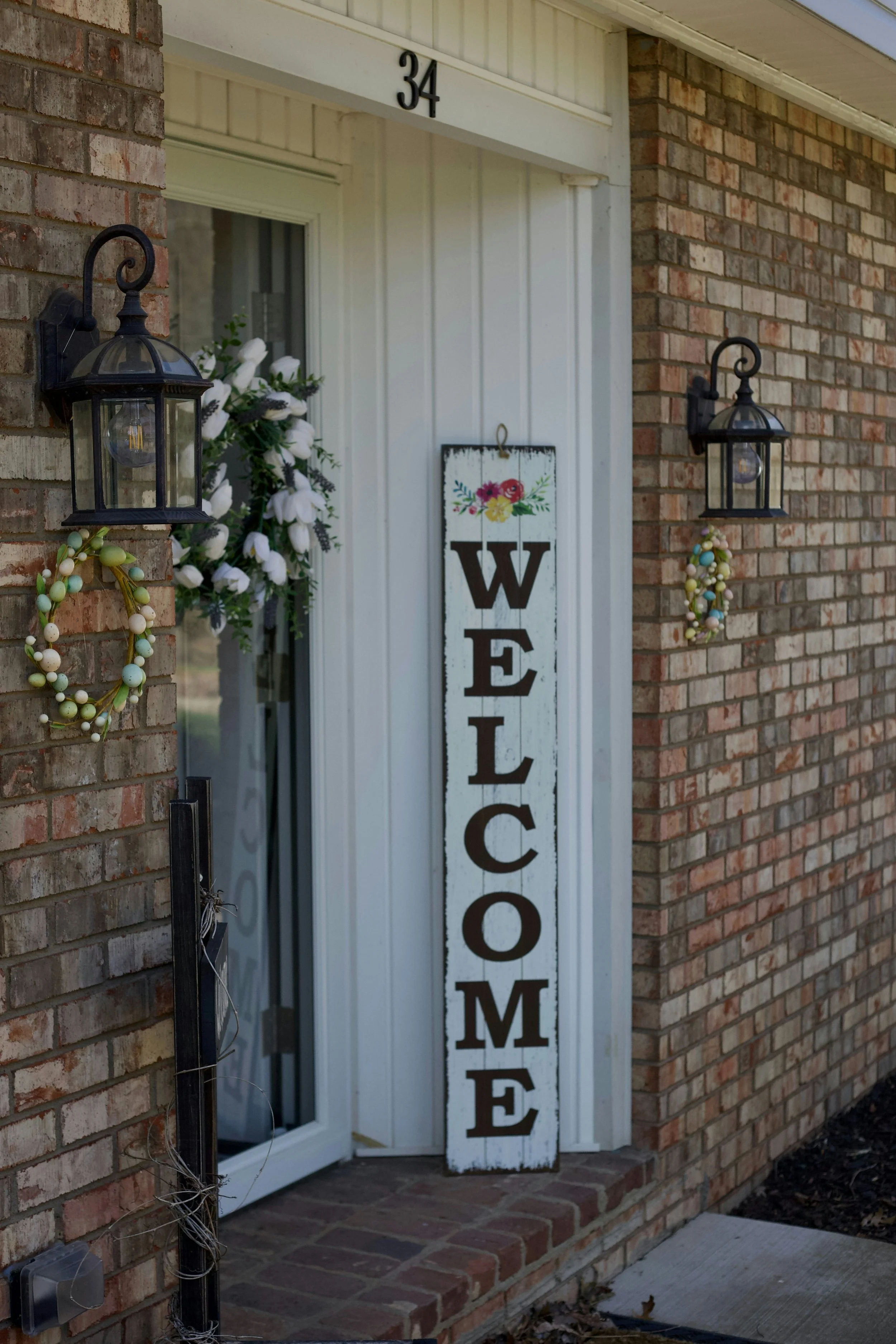 The image shows the front porch of a house with a brick exterior wall. There are two black outdoor wall lanterns mounted on the brick wall, each illuminated. A white vertical wooden sign decorated with colorful flowers and the word 'WELCOME' in large black letters is leaning against the house near the front door. There are also hanging floral and beaded decorations near the lanterns on the brick wall.