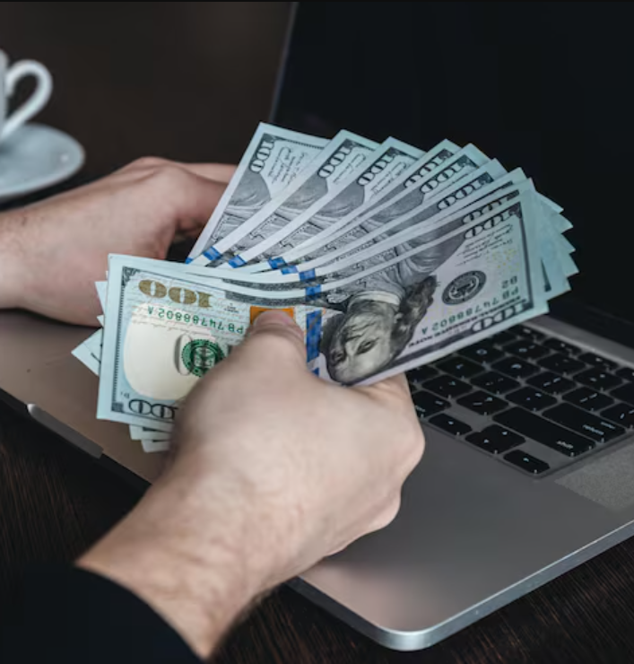 Person holding a fan of hundred-dollar bills in front of a laptop.