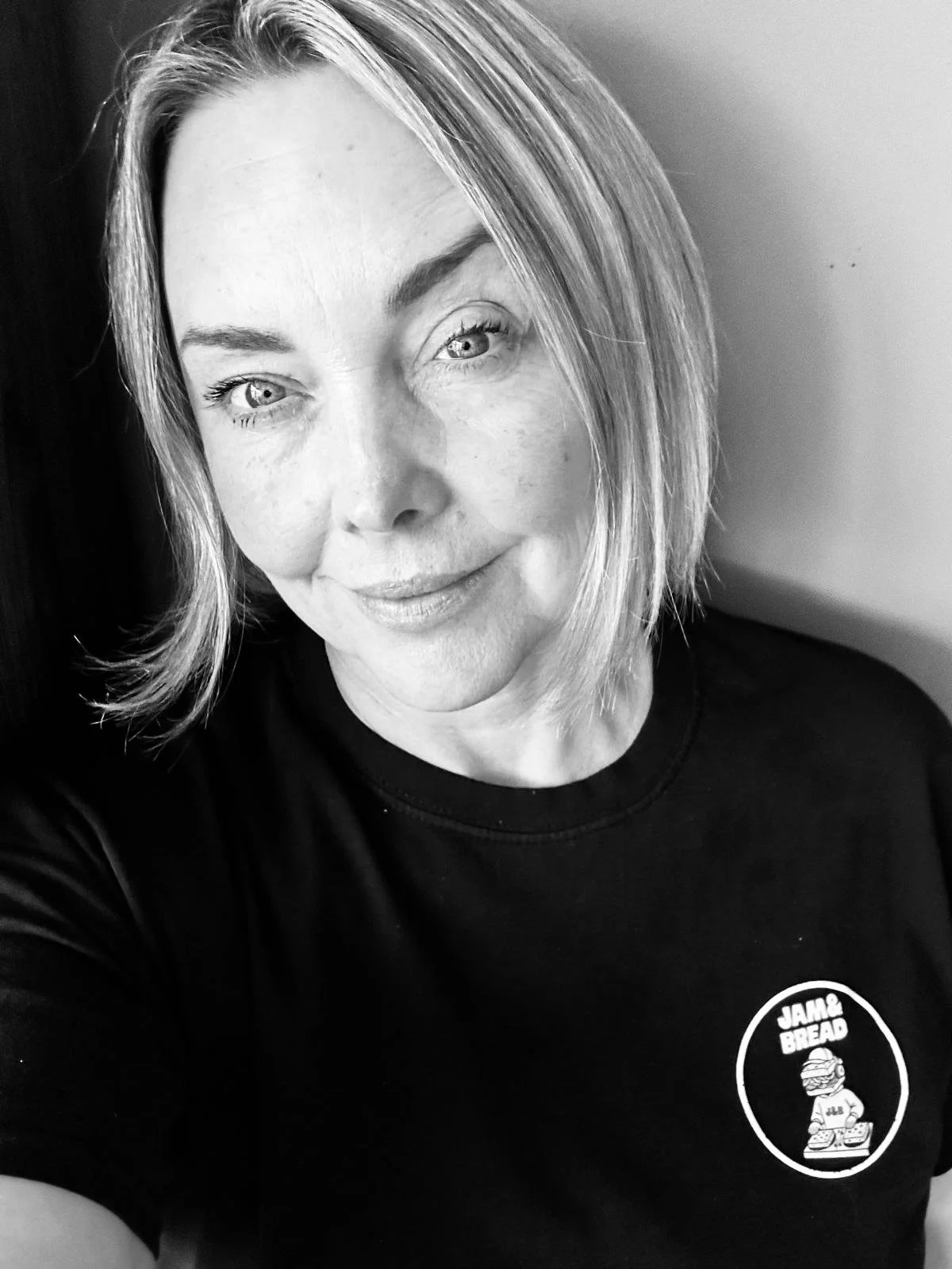 A black and white photo of a smiling woman with shoulder-length hair, wearing a T-shirt with a 'Jam & Bread' logo on it.