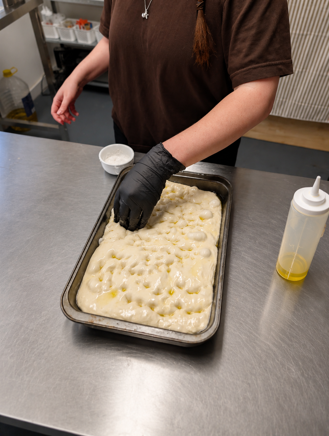 Person wearing a black glove handling a dish of uncooked cheese or dough, with olive oil and a small bowl of white liquid nearby in a kitchen or bakery setting.