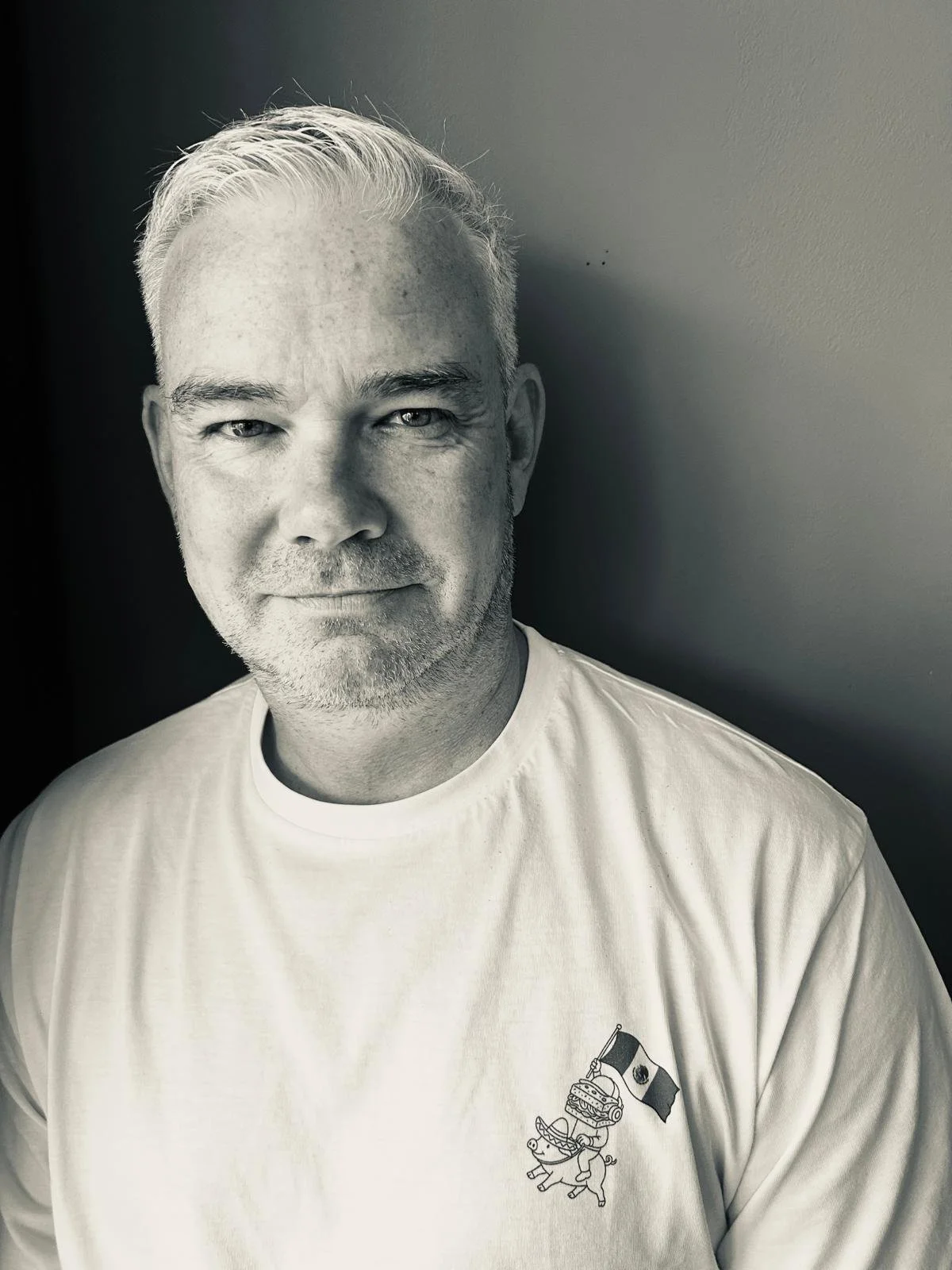 Black-and-white portrait of a middle-aged man with short, light-colored hair and a beard, smiling slightly, wearing a white t-shirt with a graphic of a pig holding a flag and a sandwich on it, against a plain background.