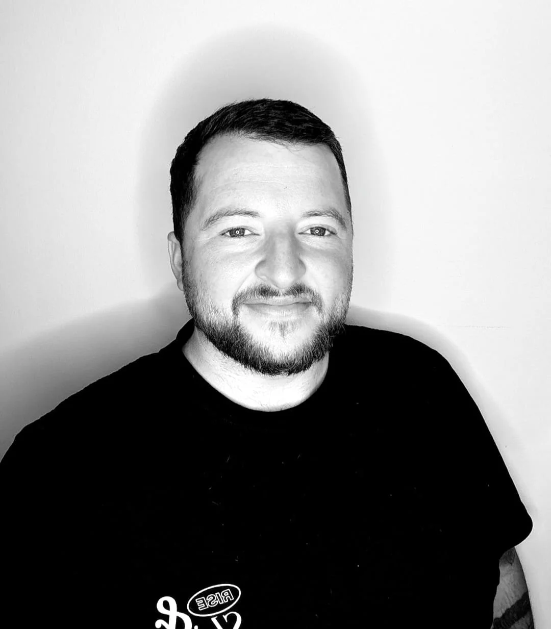 Black and white portrait of a smiling man with short dark hair and a beard, wearing a black shirt, against a plain light background.