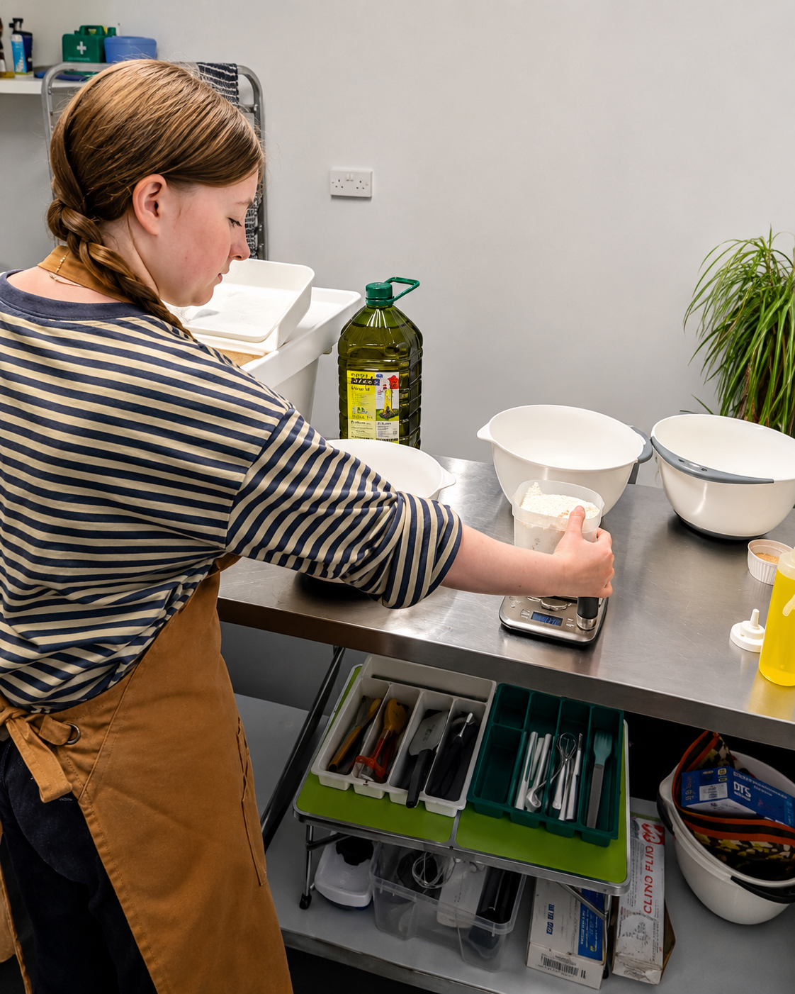 A woman wearing a striped shirt and brown apron measures flour on a digital kitchen scale in a kitchen or bakery setting. There are bowls, utensils, and cooking supplies on the stainless steel counter and shelves behind her.