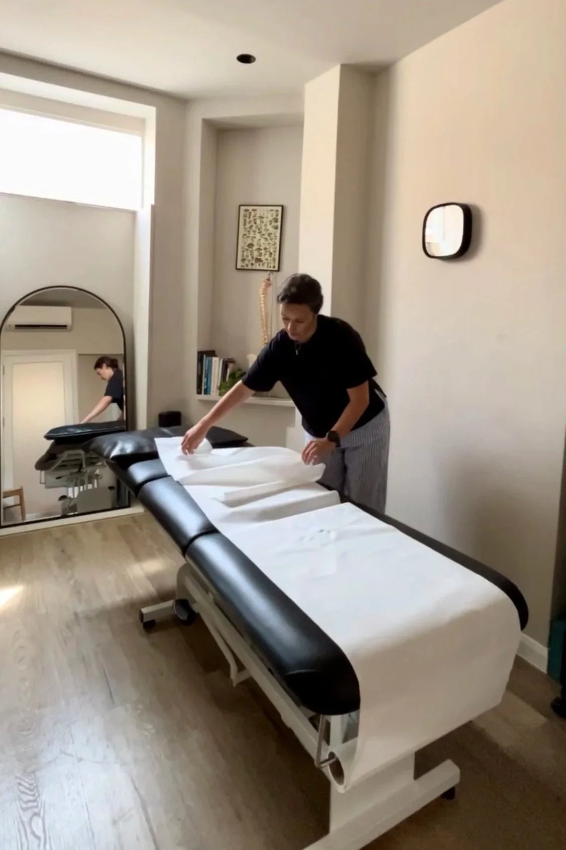 A person making a bed in a room that appears to be a medical or massage therapy studio, with a treatment table, a large mirror, and a bookshelf on the wall.